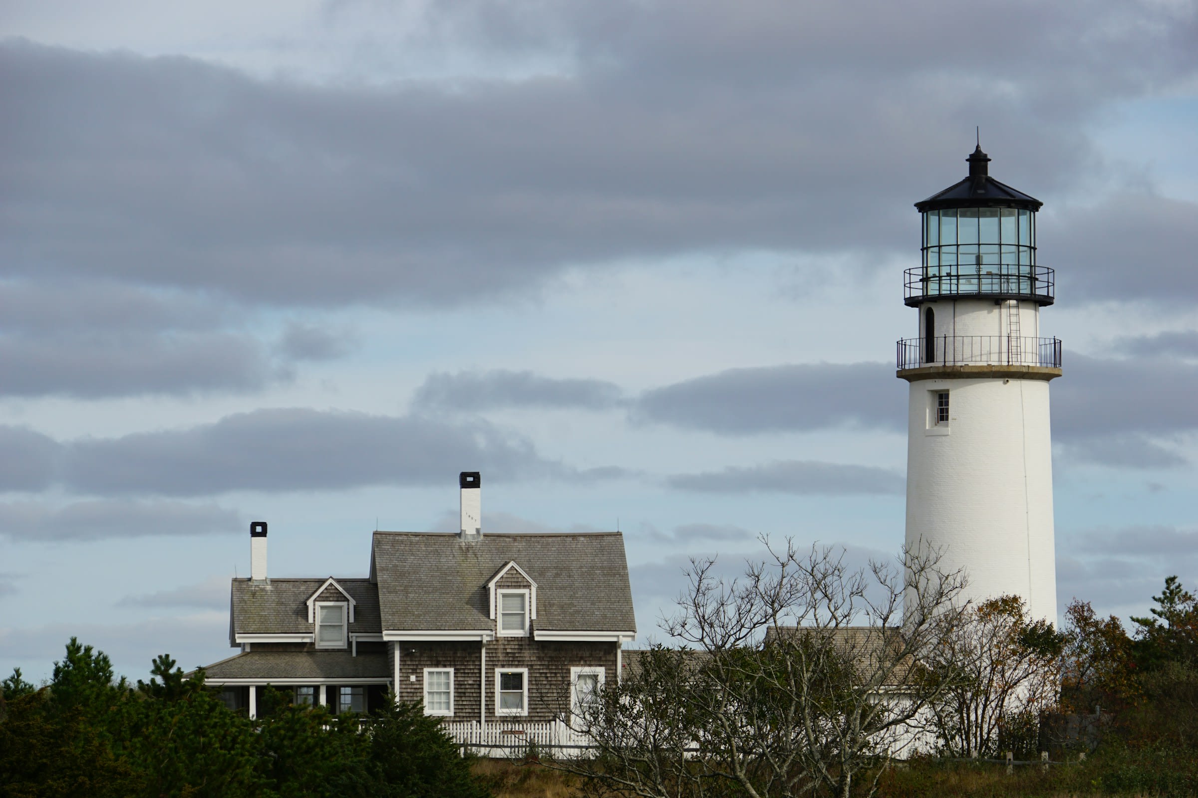a white lighthouse and a grey shingle house on a cloudy day