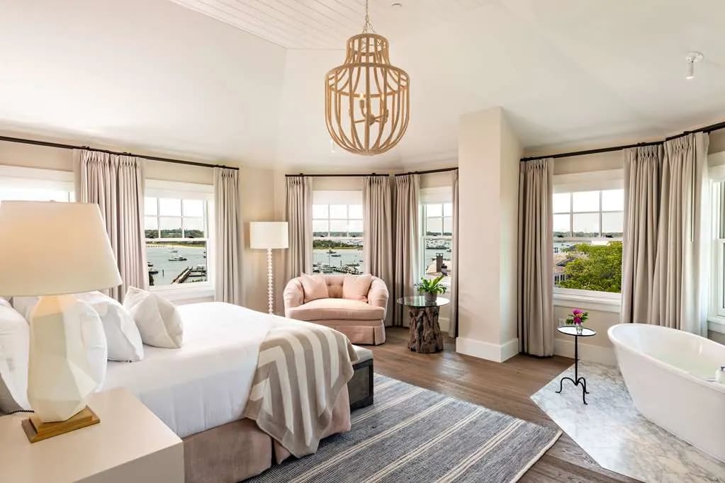 A neutral coastal chic style hotel room with white walls, tan drapes, light wood chandelier, grey and white striped carpet and blanket.