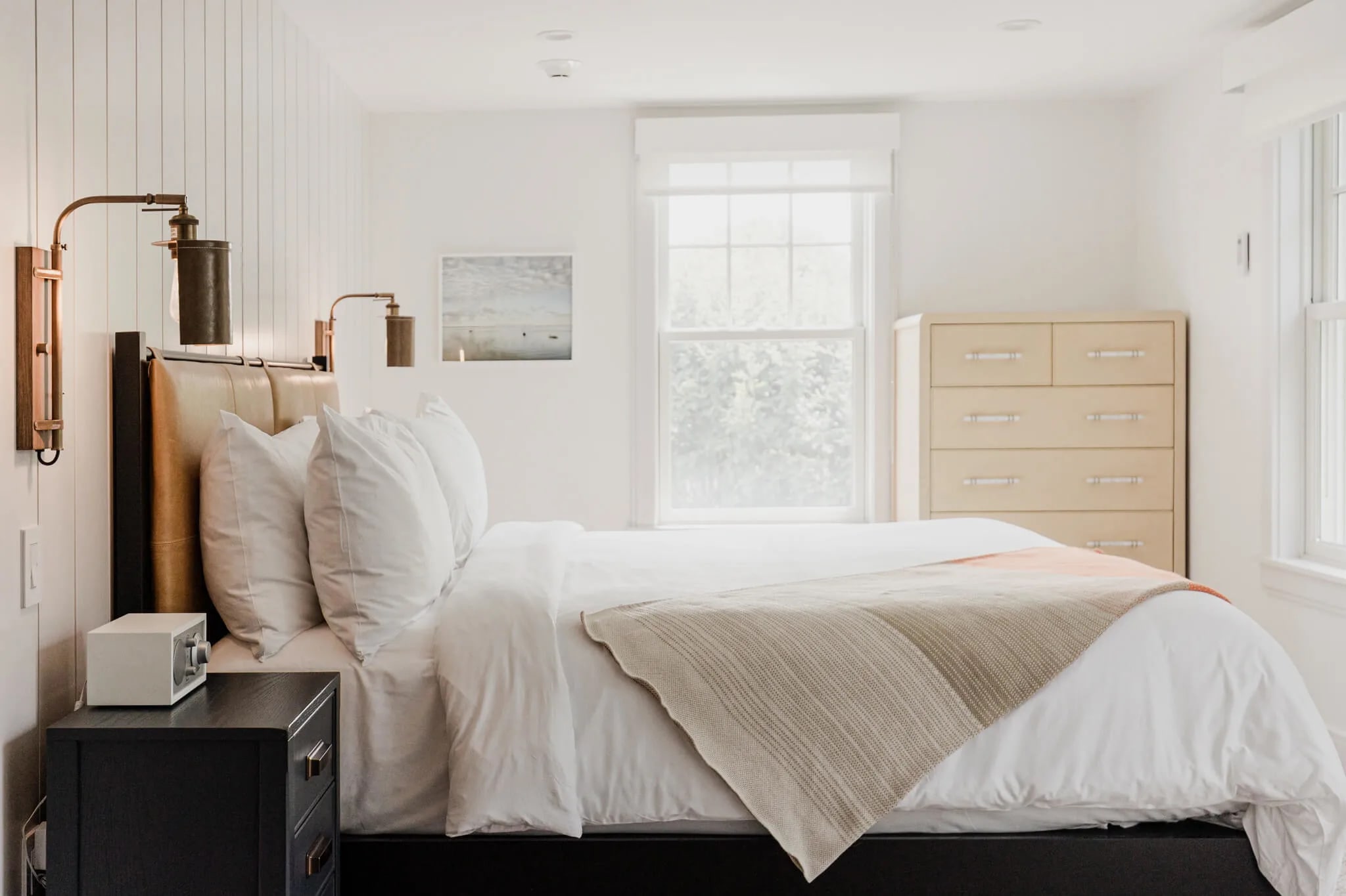 A small white hotel room with light wood bureau, black night table and headboard, with white shiplap wall and white linens