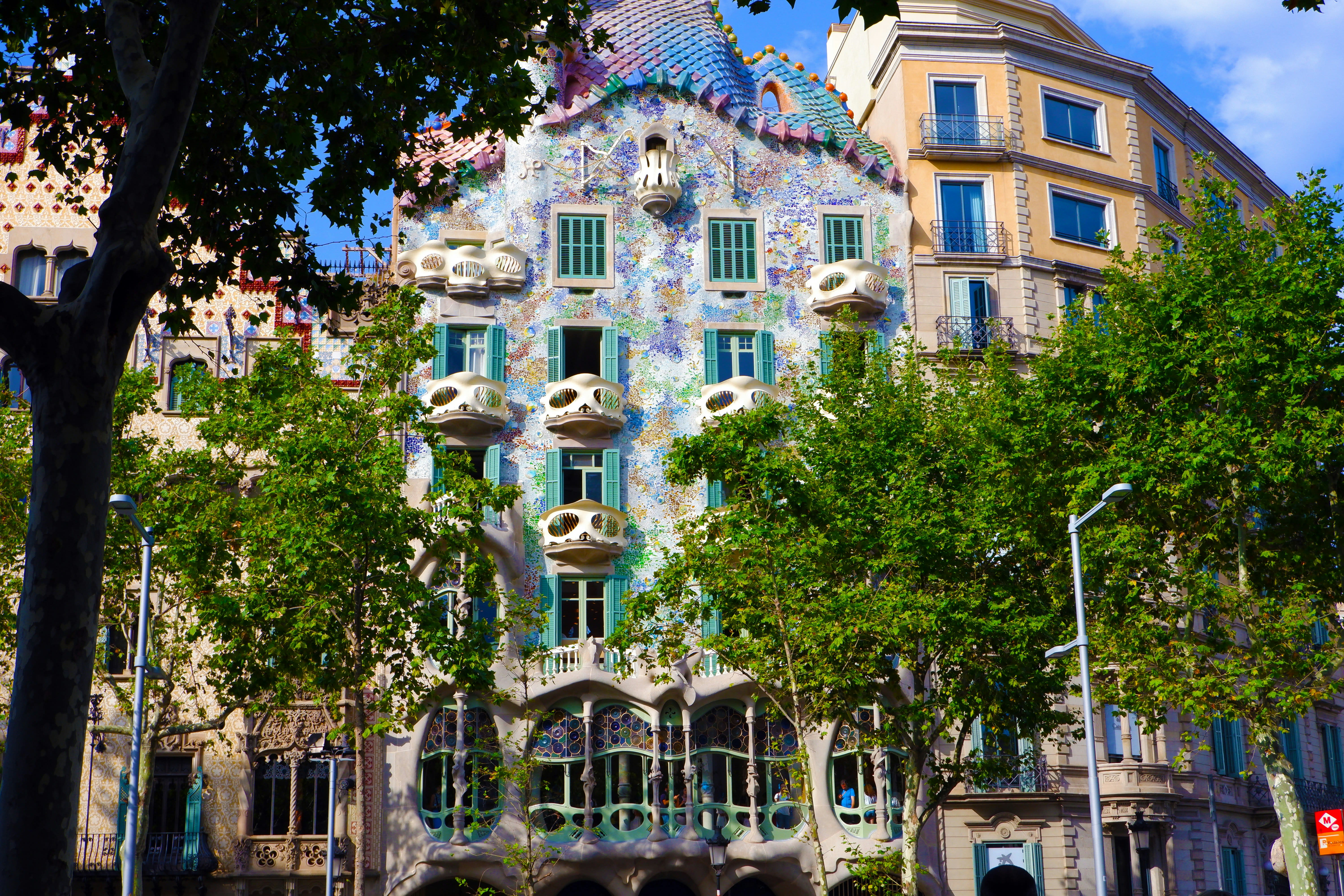 A colourful, mosaic-covered building with wavy lines and bone-like balconies, partially obscured by leafy trees.