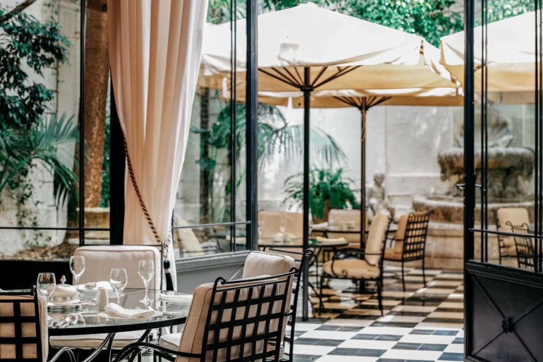 An indoor and outdoor restaurant with cream umbrellas, checkered floor tiles and greenery.