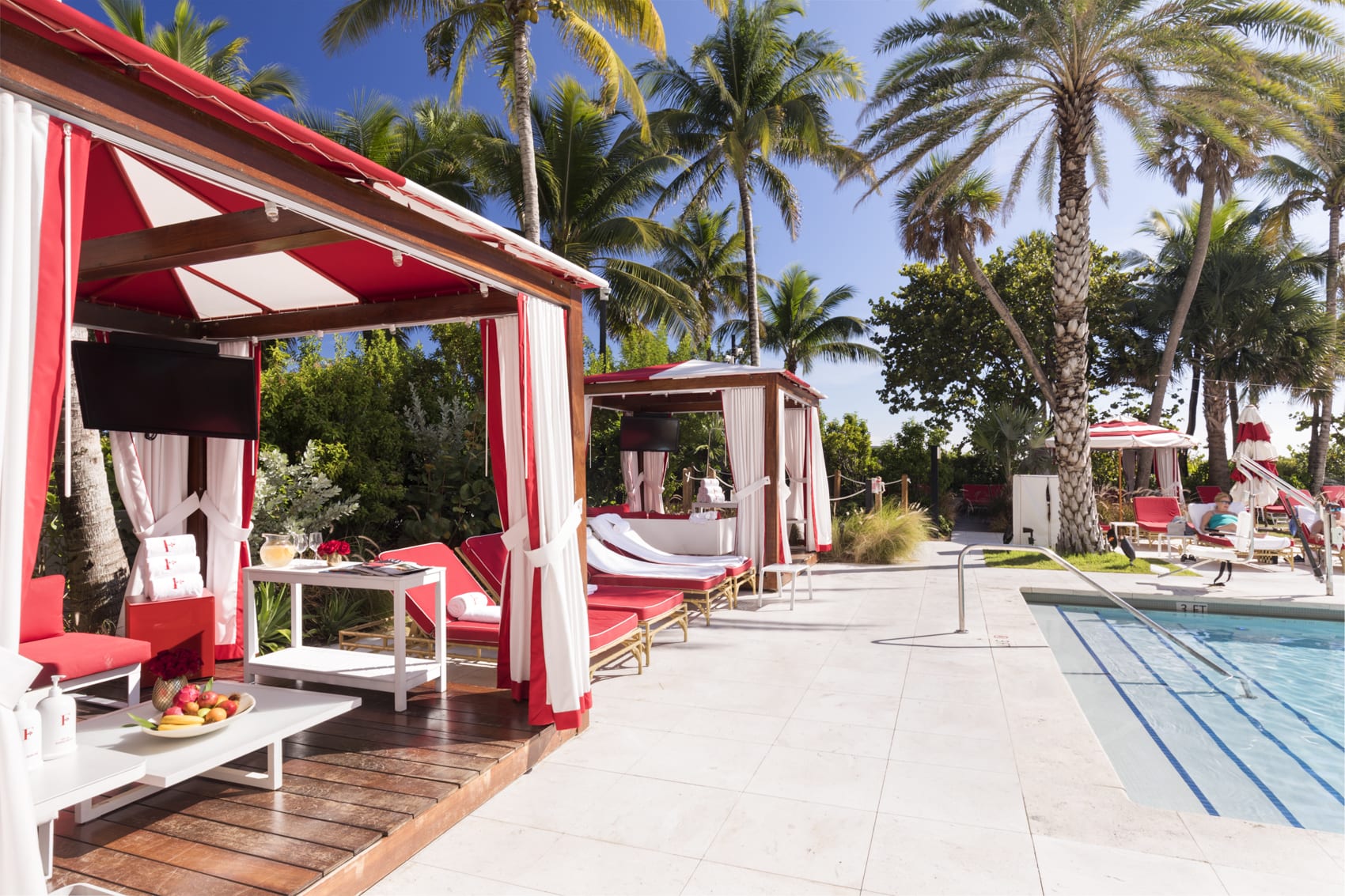 A pool area with surrounding palm trees and red-and-white striped cabanas and loungers.