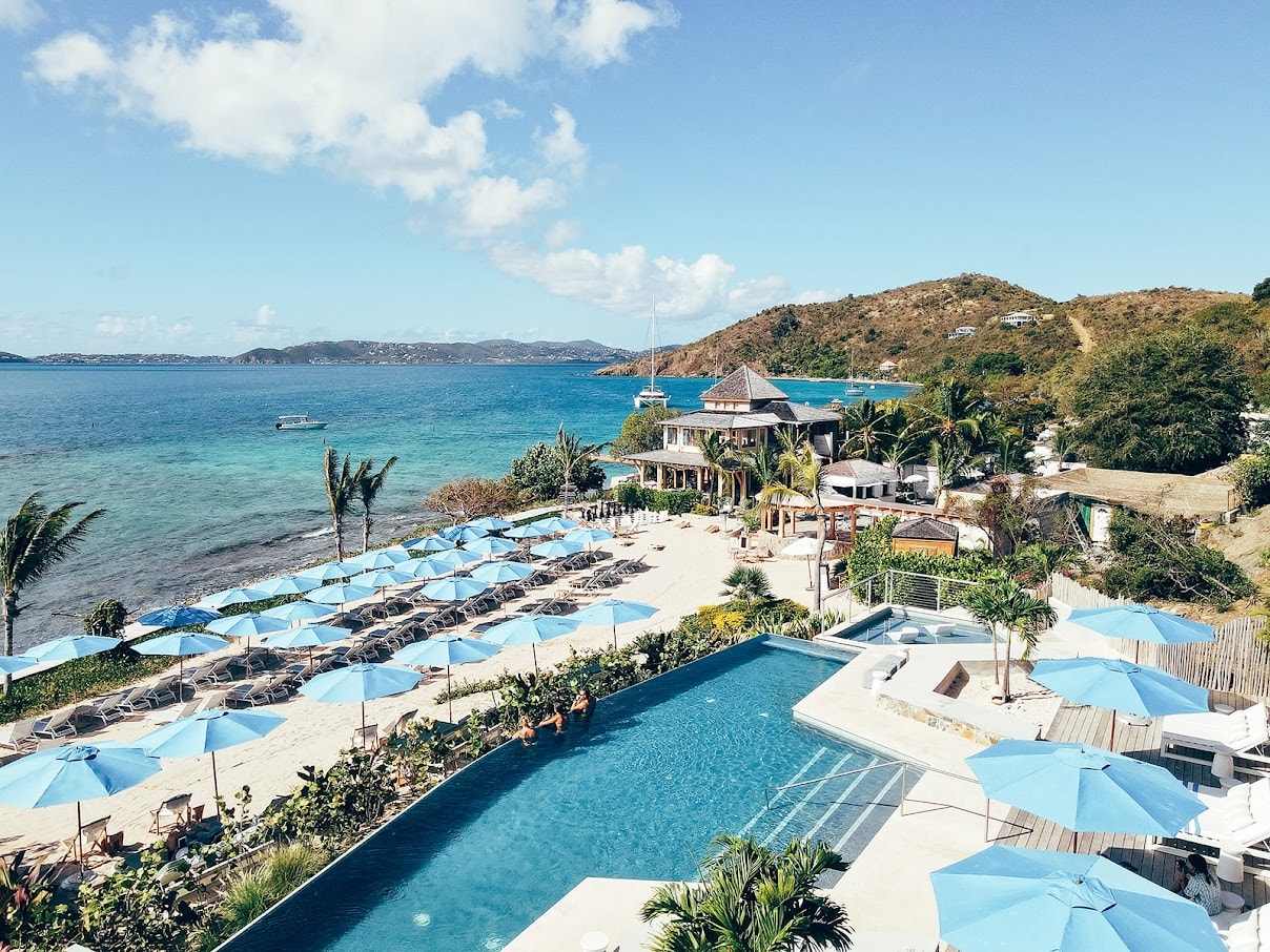 An aerial view of a pool area leading down to a tropical beach with light blue umbrellas and loungers.