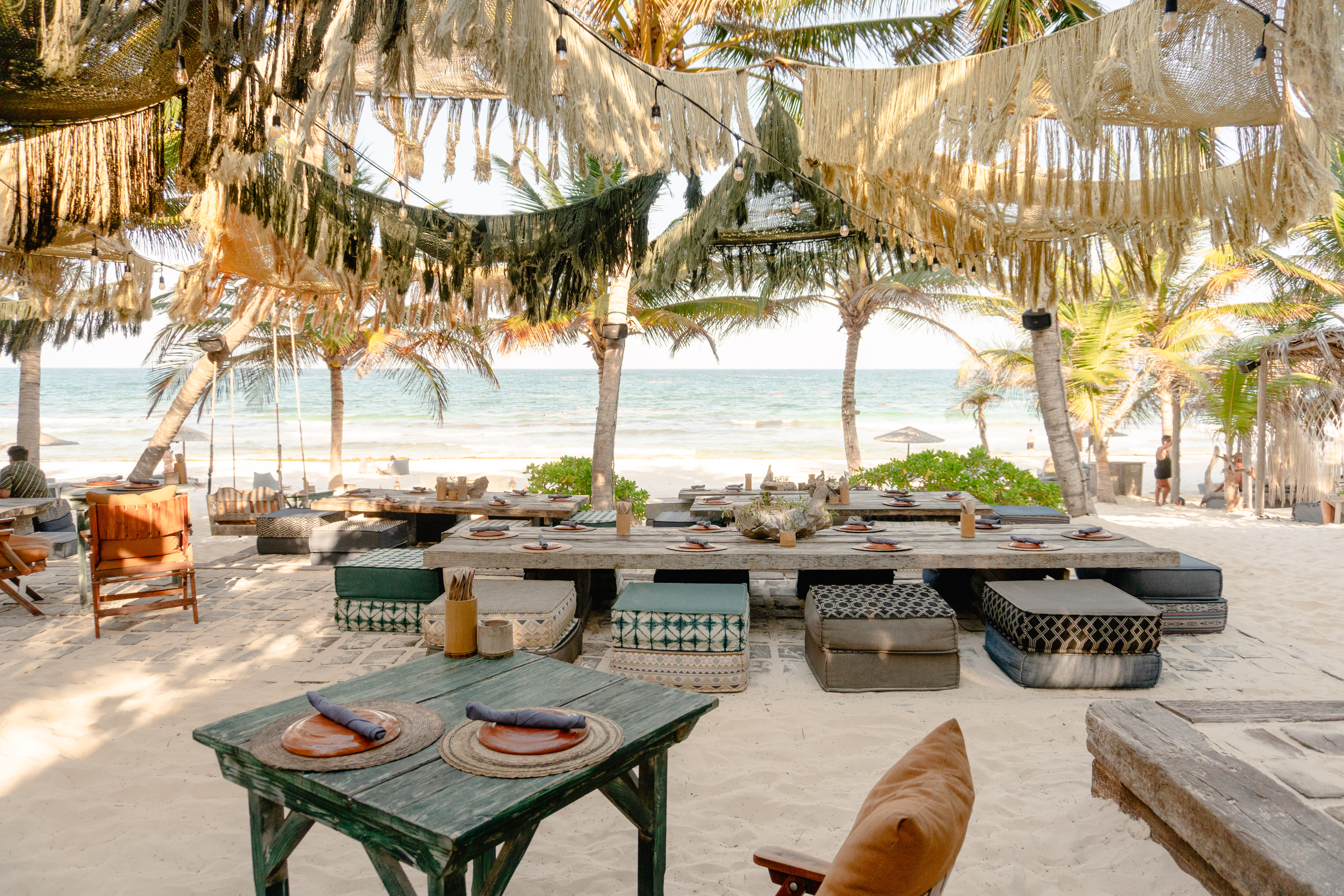 A beachfront restaurant with low wooden tables, cushioned seating, and boho-style fringe canopies overlooking the ocean.