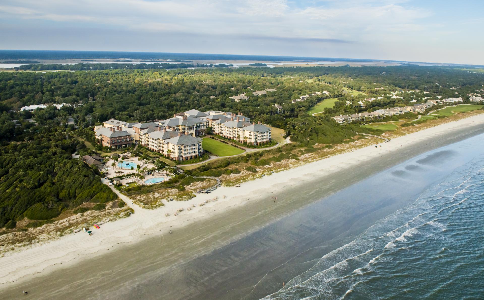 An aerial view of a large, beachfront resort, surrounded by greenery and rural landscape