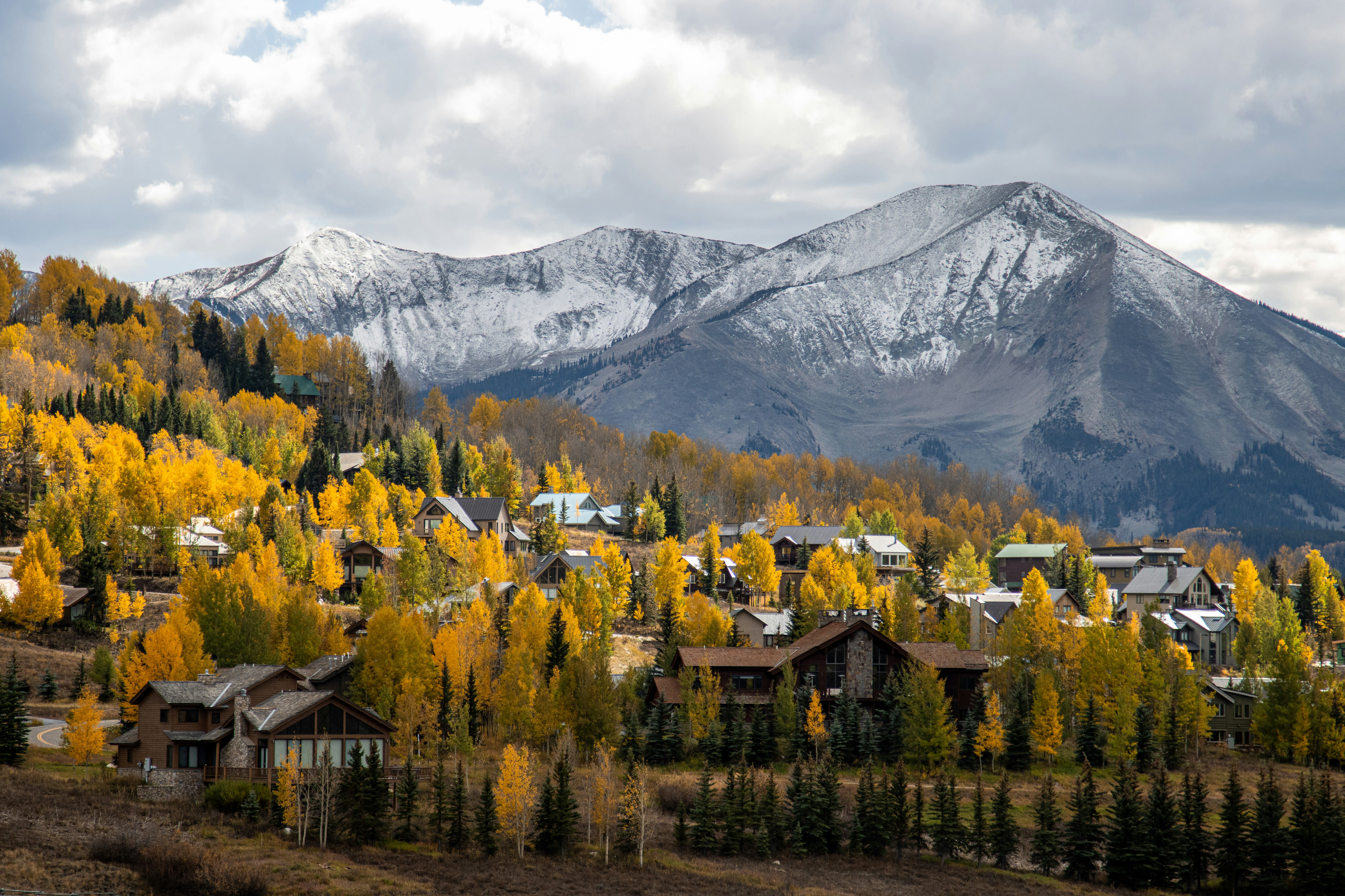 Snowy mountain peaks surrounded by fall foliage with scattered buildings.