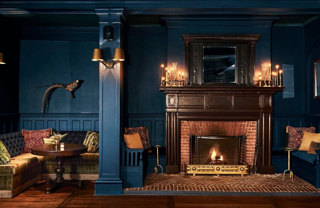 A cozy lounge area with dark blue walls and pillars, warm lighting and a crackling fireplace.