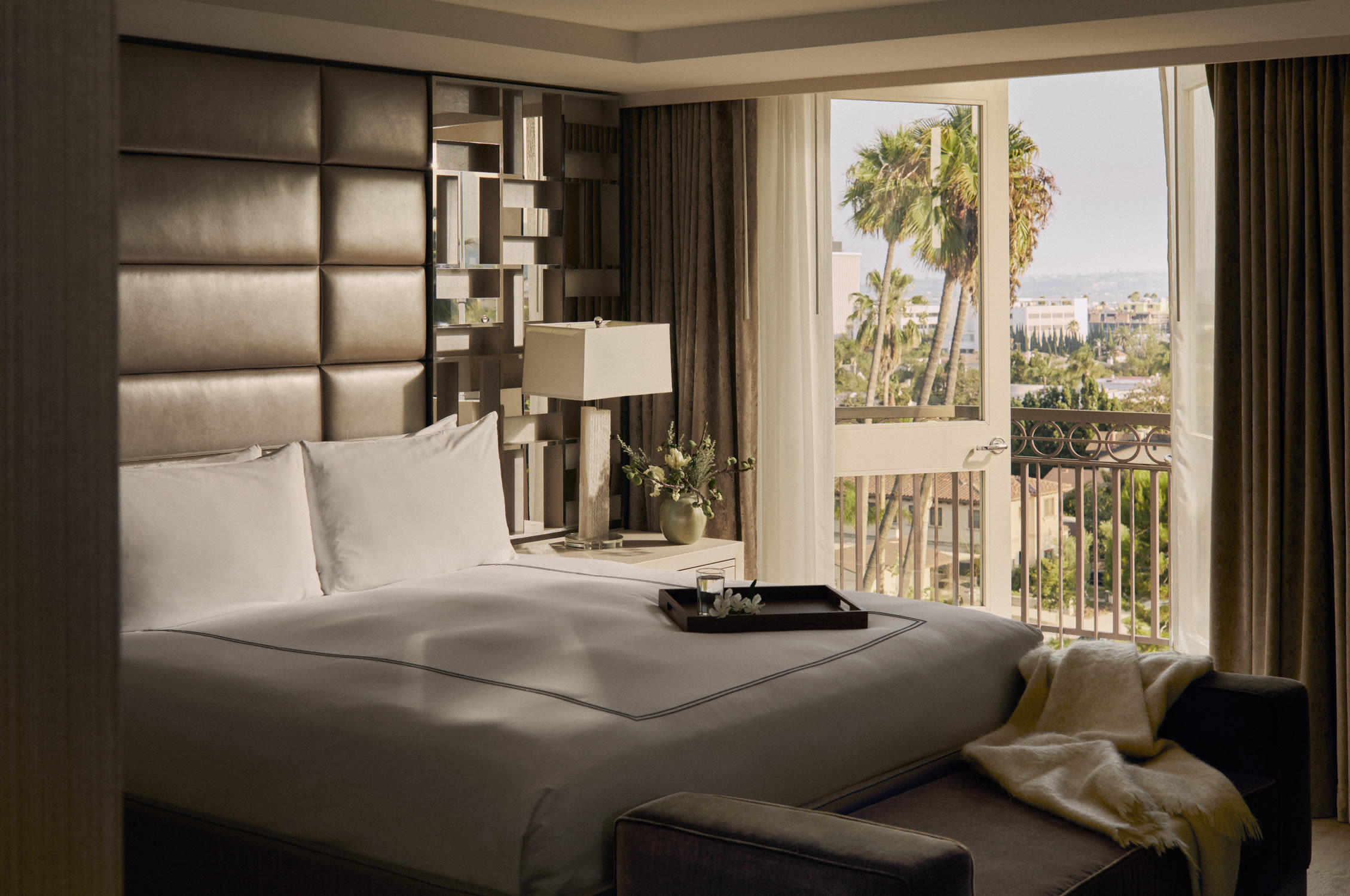 An elegant hotel room with padded headboard and white bedding in dim light, and an open balcony door leading out to a view with palm treets.