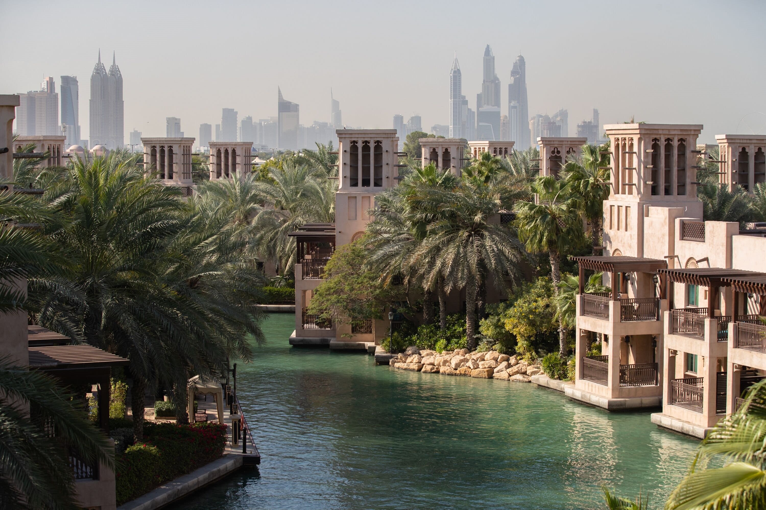 Arabian heritage architecture homes with a lake and palm trees, and skyscrapers on the horizon behind them