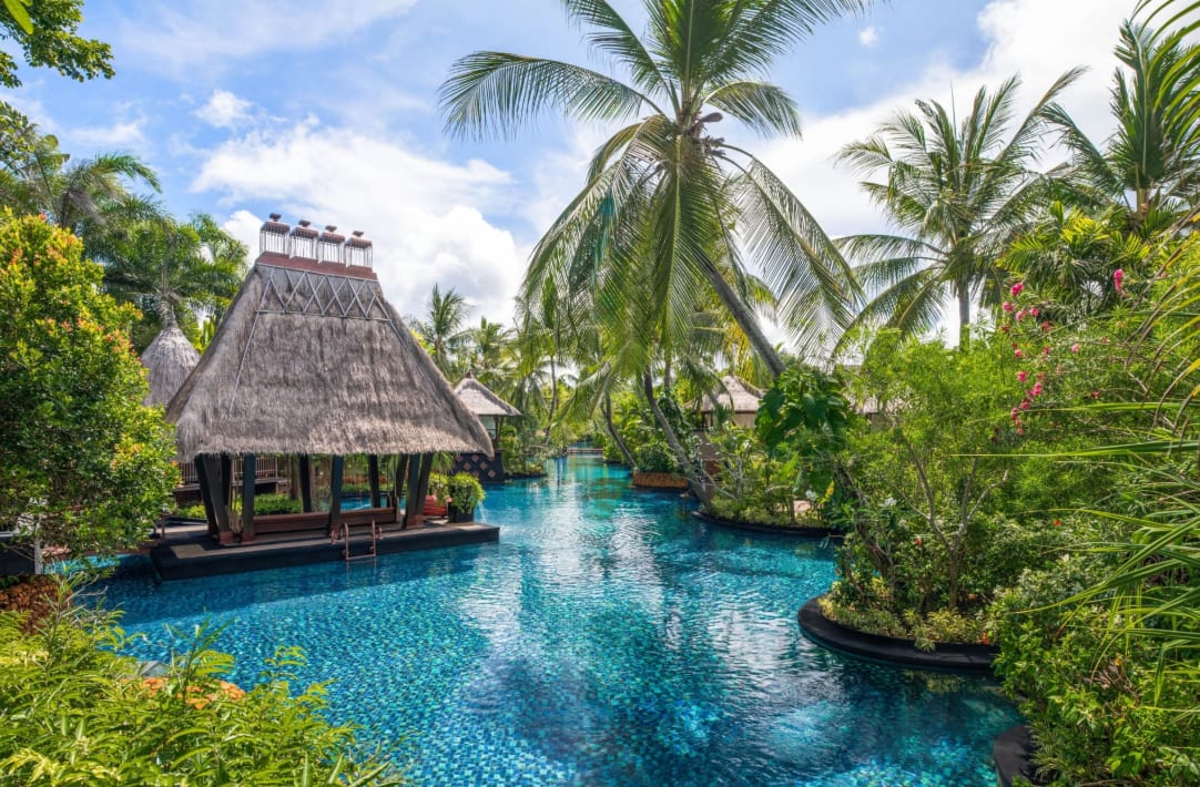 A lagoon area at a hotel with tropical foliage and a building with a straw roof.