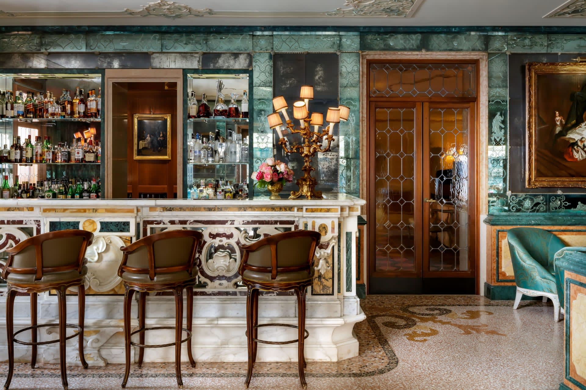 An ornate Italian bar with white marble bar, green marble walls, alcohol on glass shelves and wood chairs