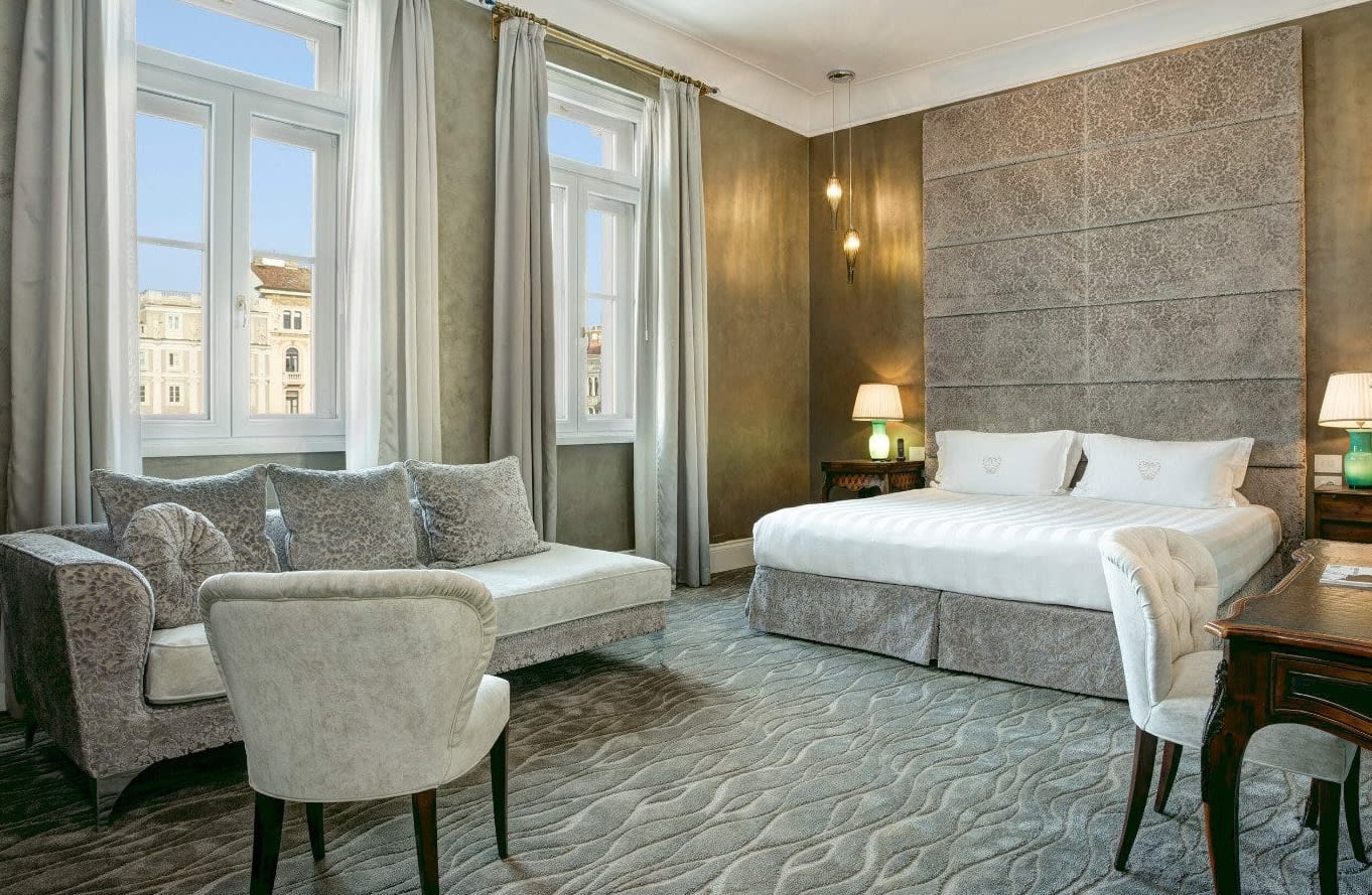 A modern hotel room with grey carpet, grey sofa, grey cloth headboard, gold walls and white linen sheets.