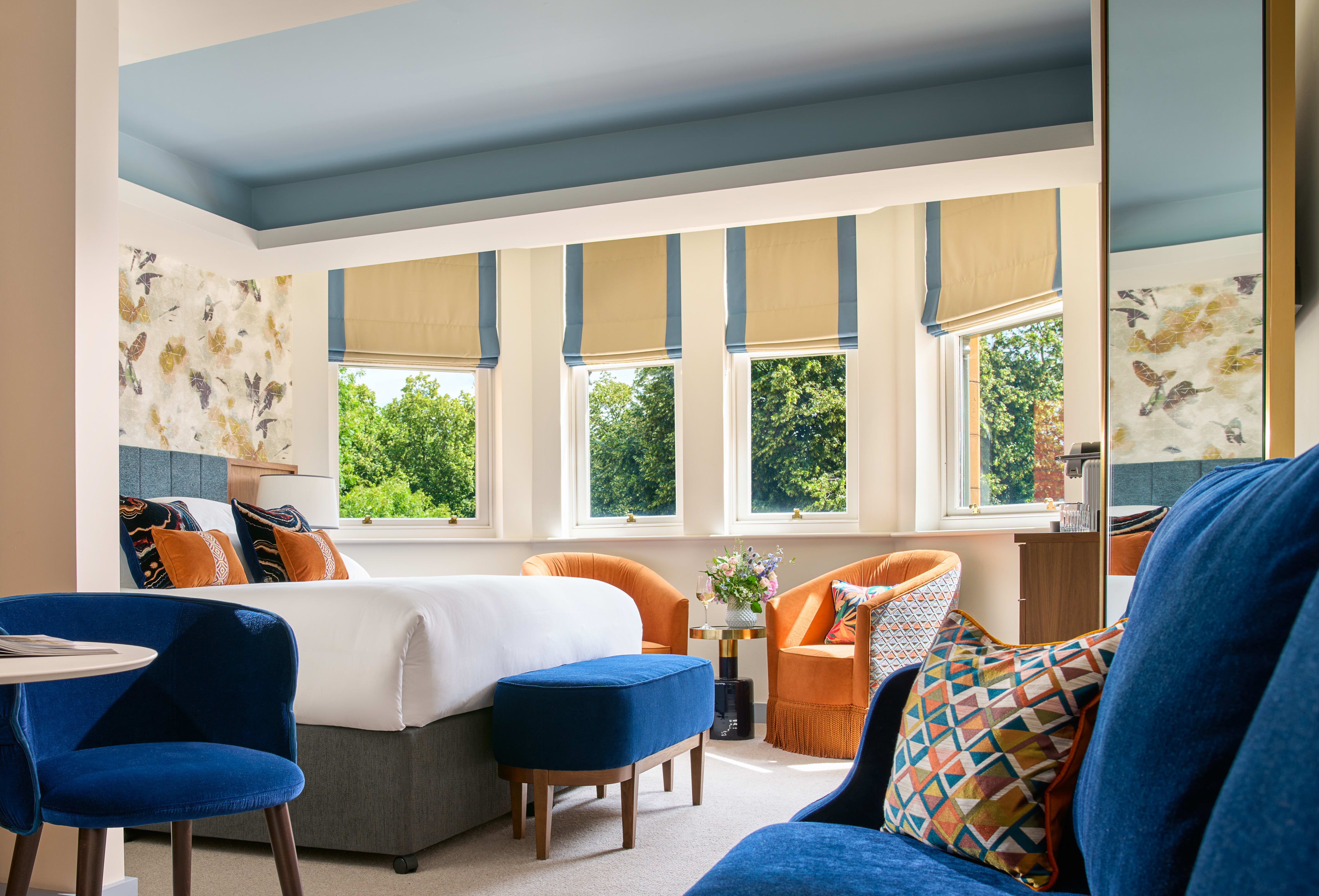 A bright, colorful hotel room with blue and orange accents and furnishing, with a view of greenery through windows