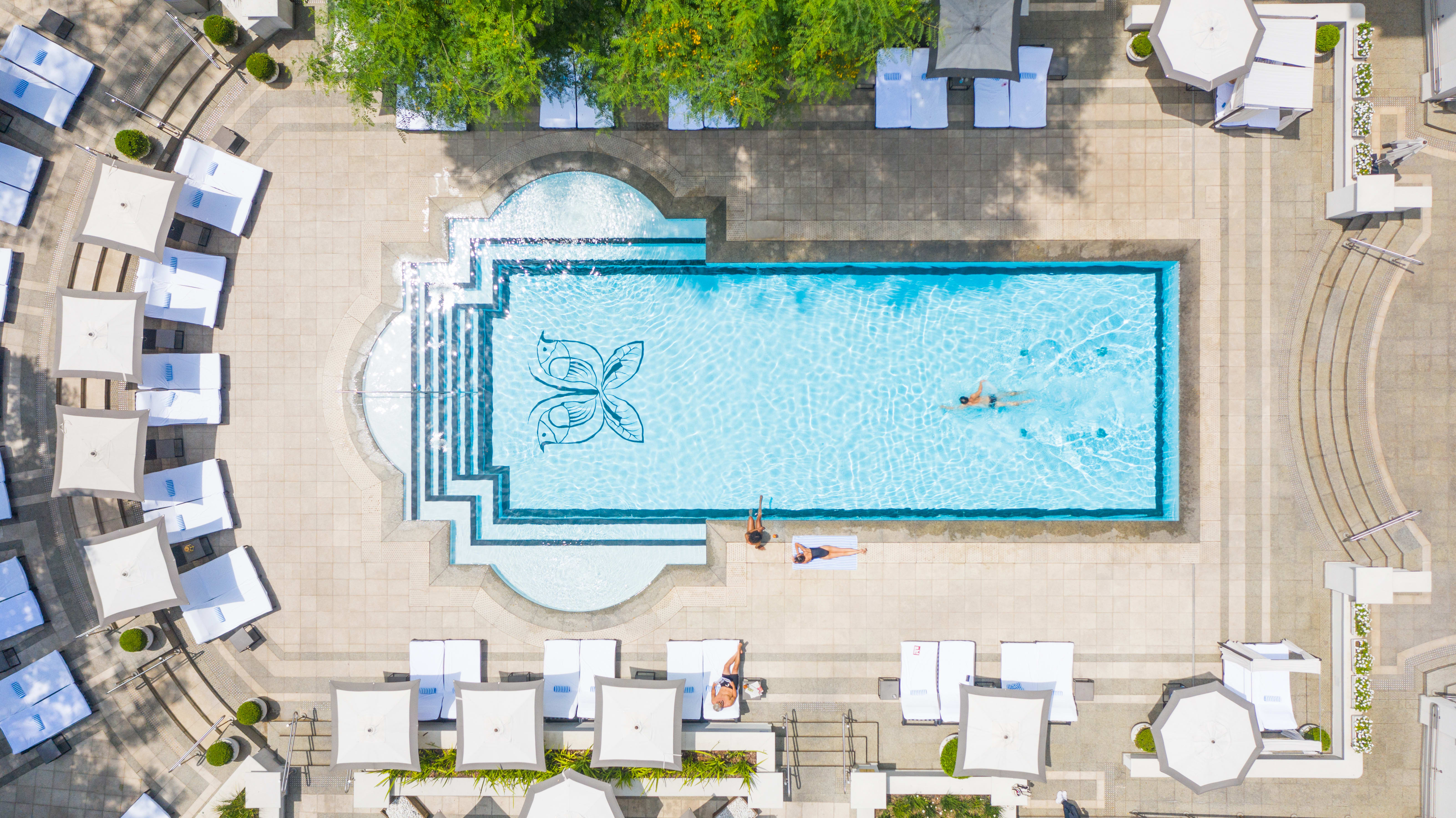 An aerial image of an elegant outdoor hotel pool with logo printed on the base of the pool with white sun loungers and greenery surrounding pool area.