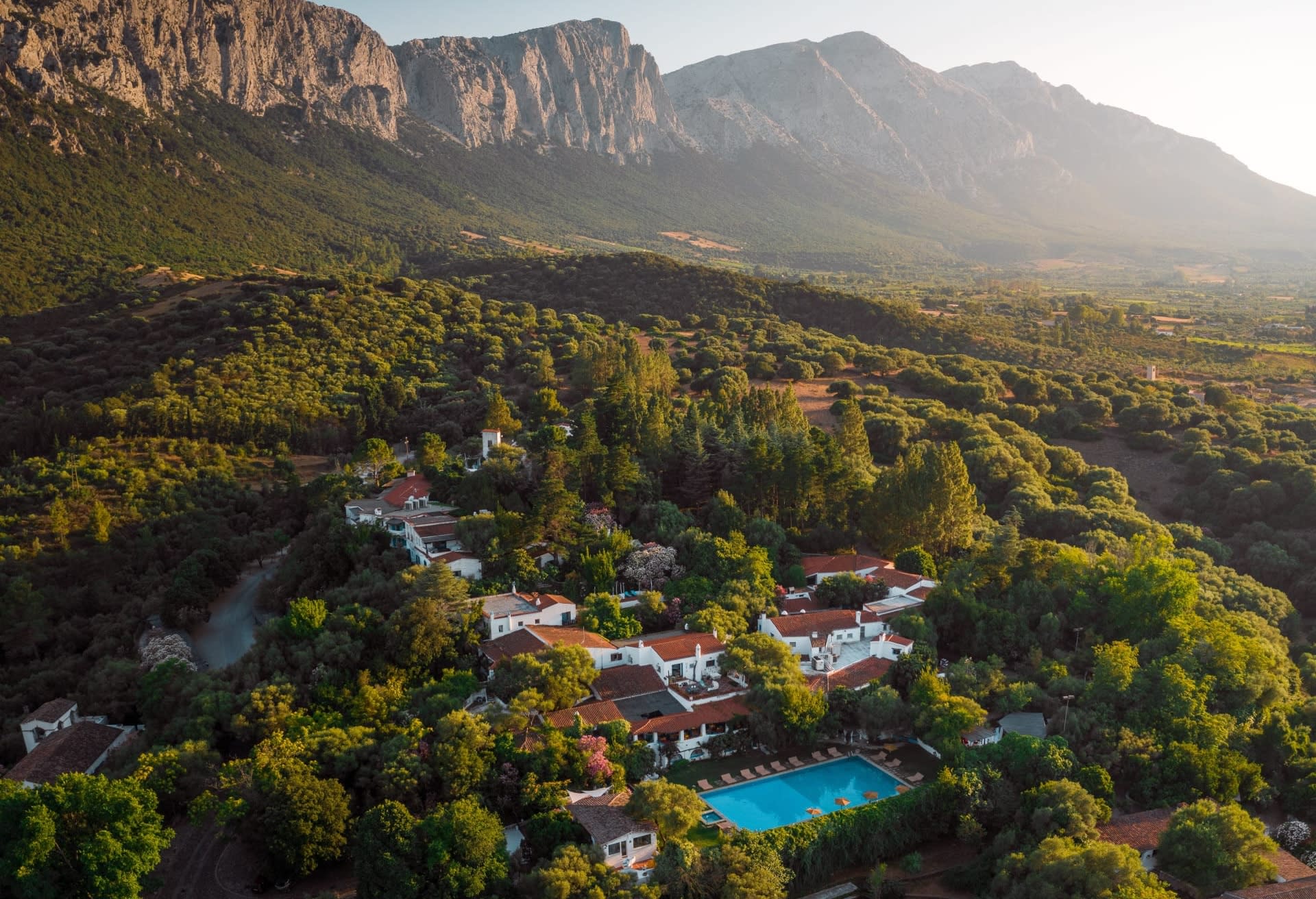 Aerial view of white buildings and pool amidst a forest and rocky mountains in the background during day
