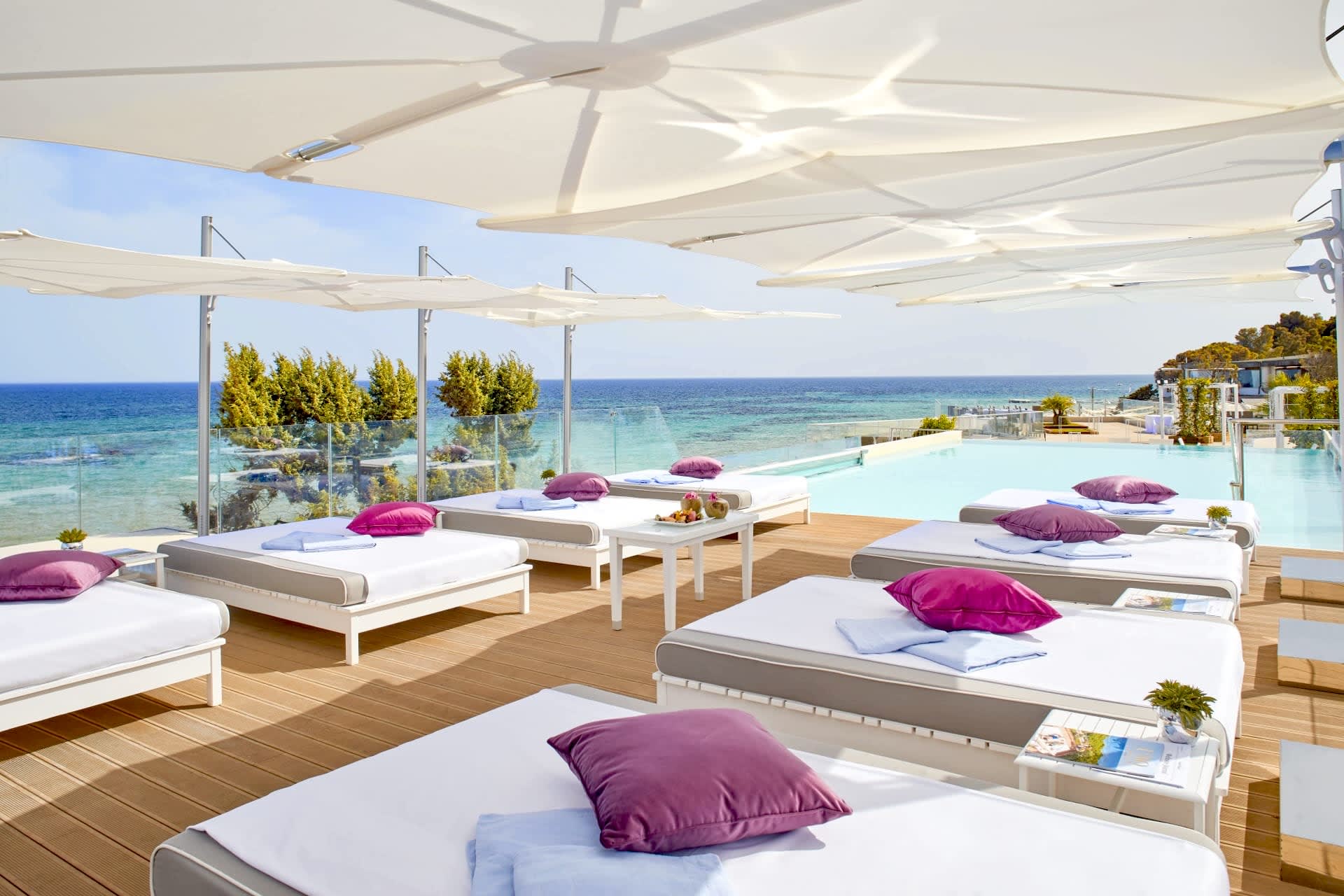 A poolside beach club with wood floors, white day beds and purple pillows, with turquoise ocean in the background during day
