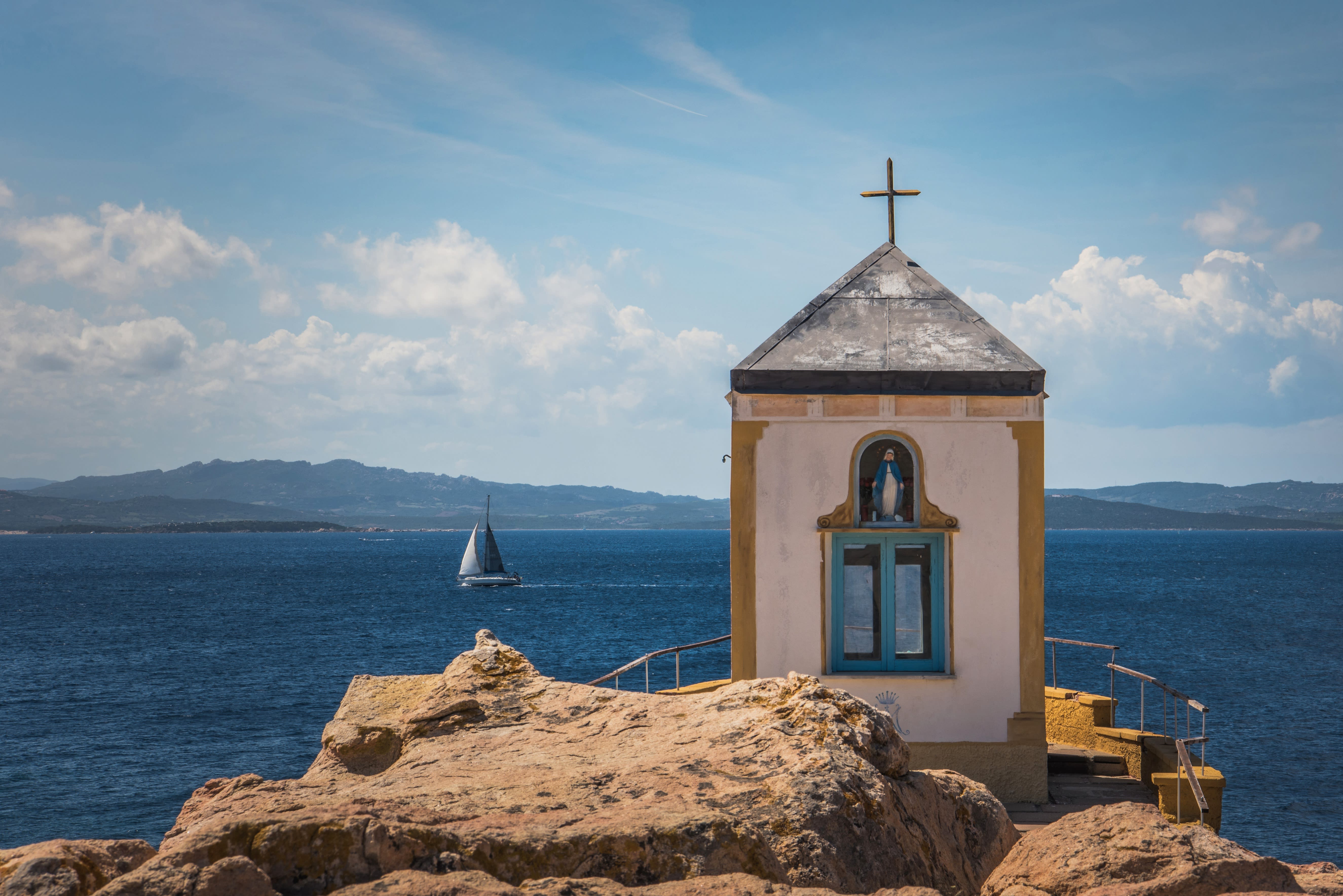 A religious building with cross on top on rocks with the ocean in the background and a sailboat during day