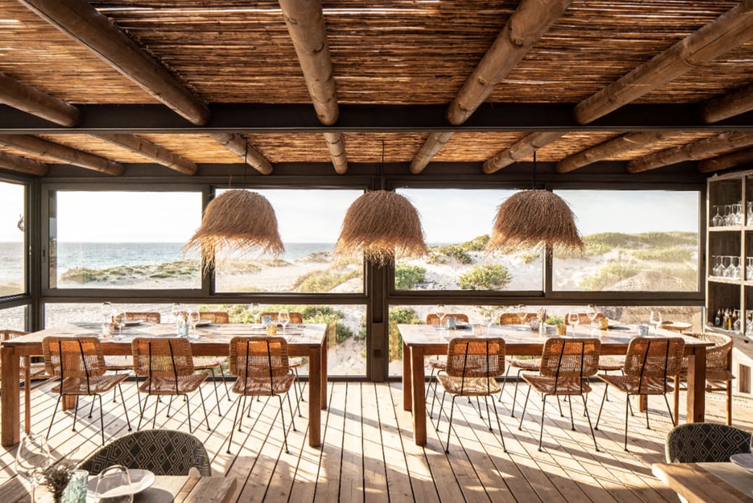 A beach club restaurant with a boho-chic design, including hanging straw lamps and a wooden deck overlooking a wild beach.