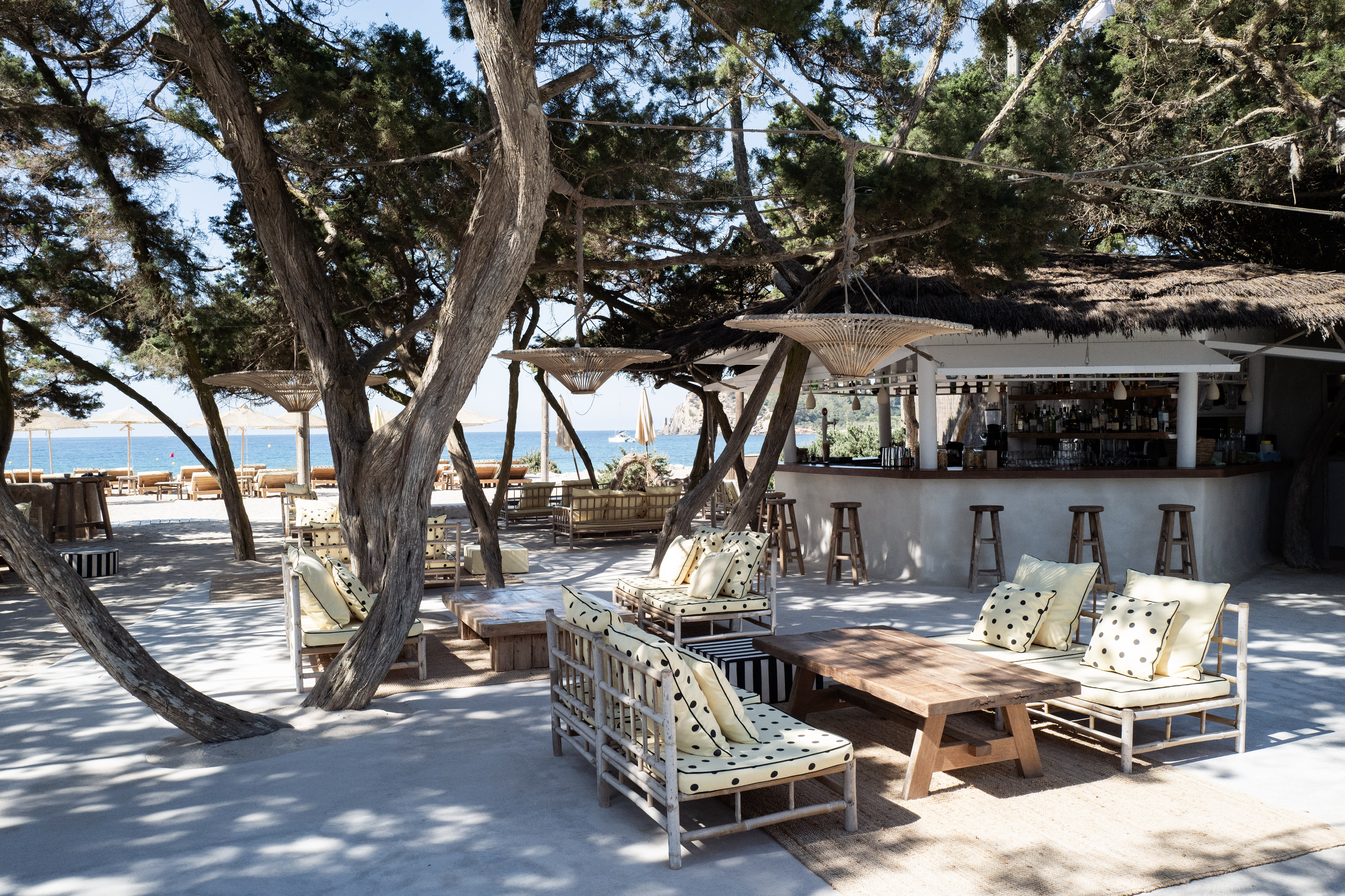 Shaded outdoor beach lounge with bamboo sofas, wooden tables, a bar, and a view of the sea.