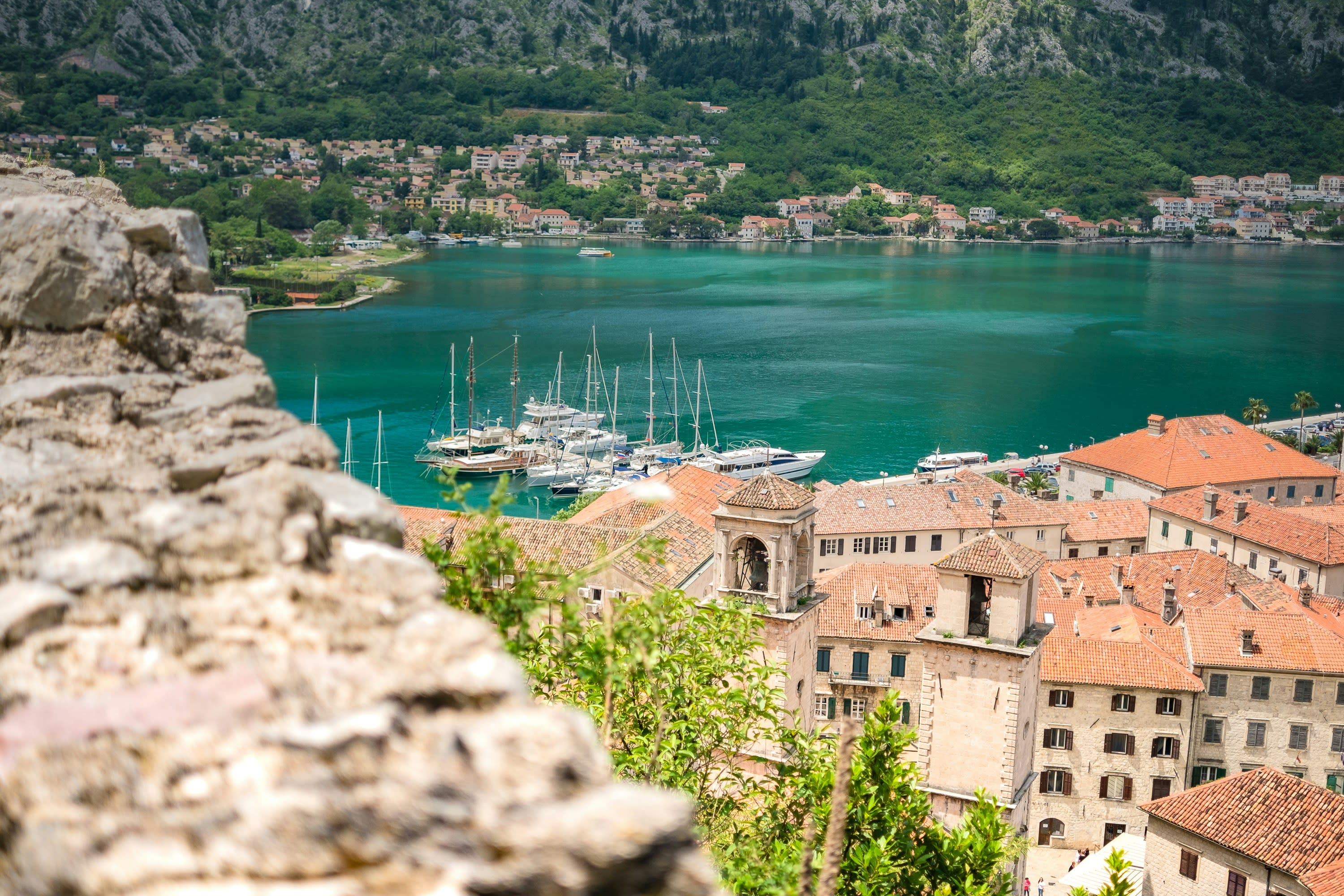 Aerial view of a coastal town with terracotta-roofed buildings, a marina with sailboats, and green hills in the background.