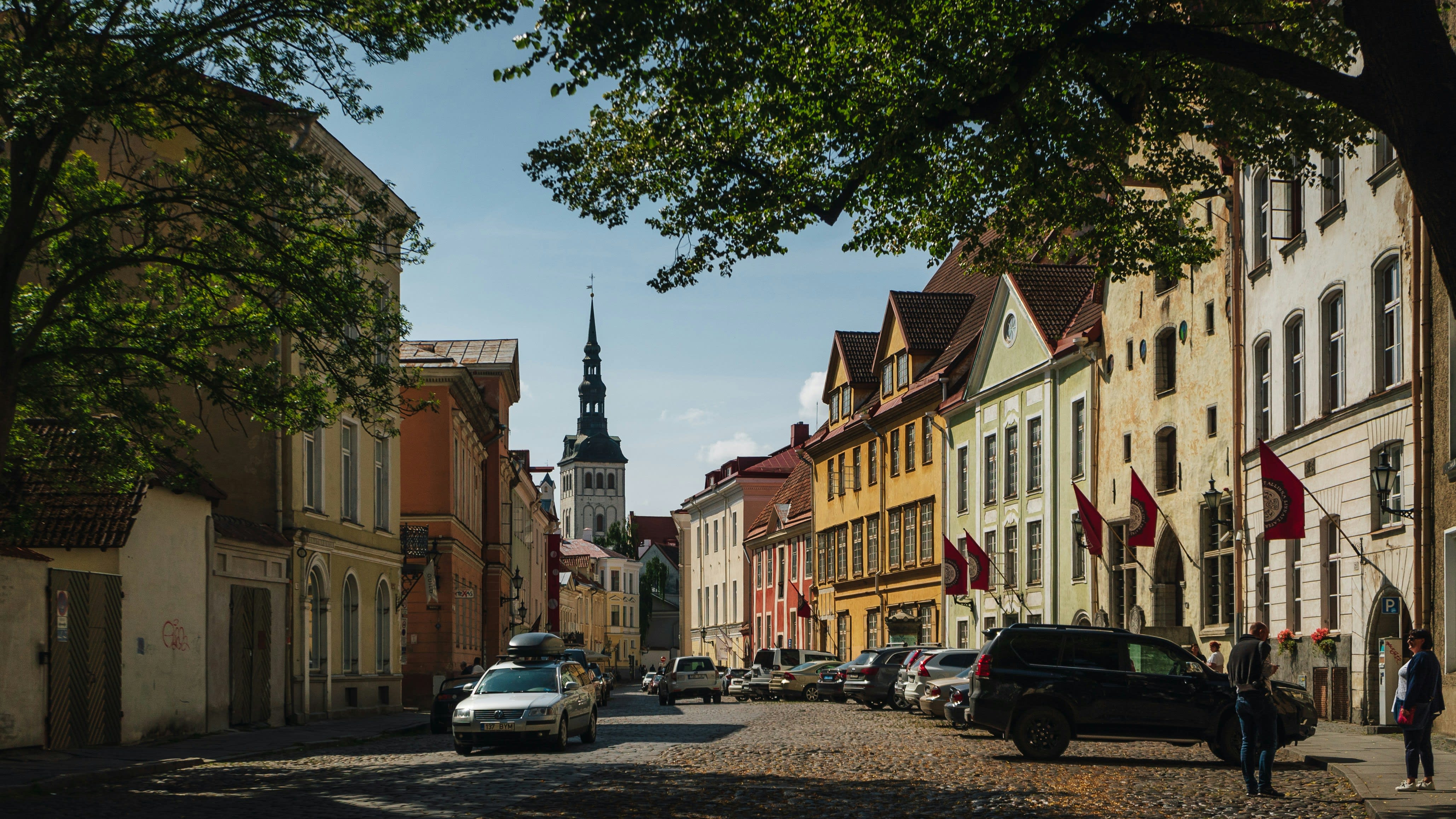 Cobblestone street in a historic European town lined with colorful old buildings, cars parked along the side, and a church spire in the background.
