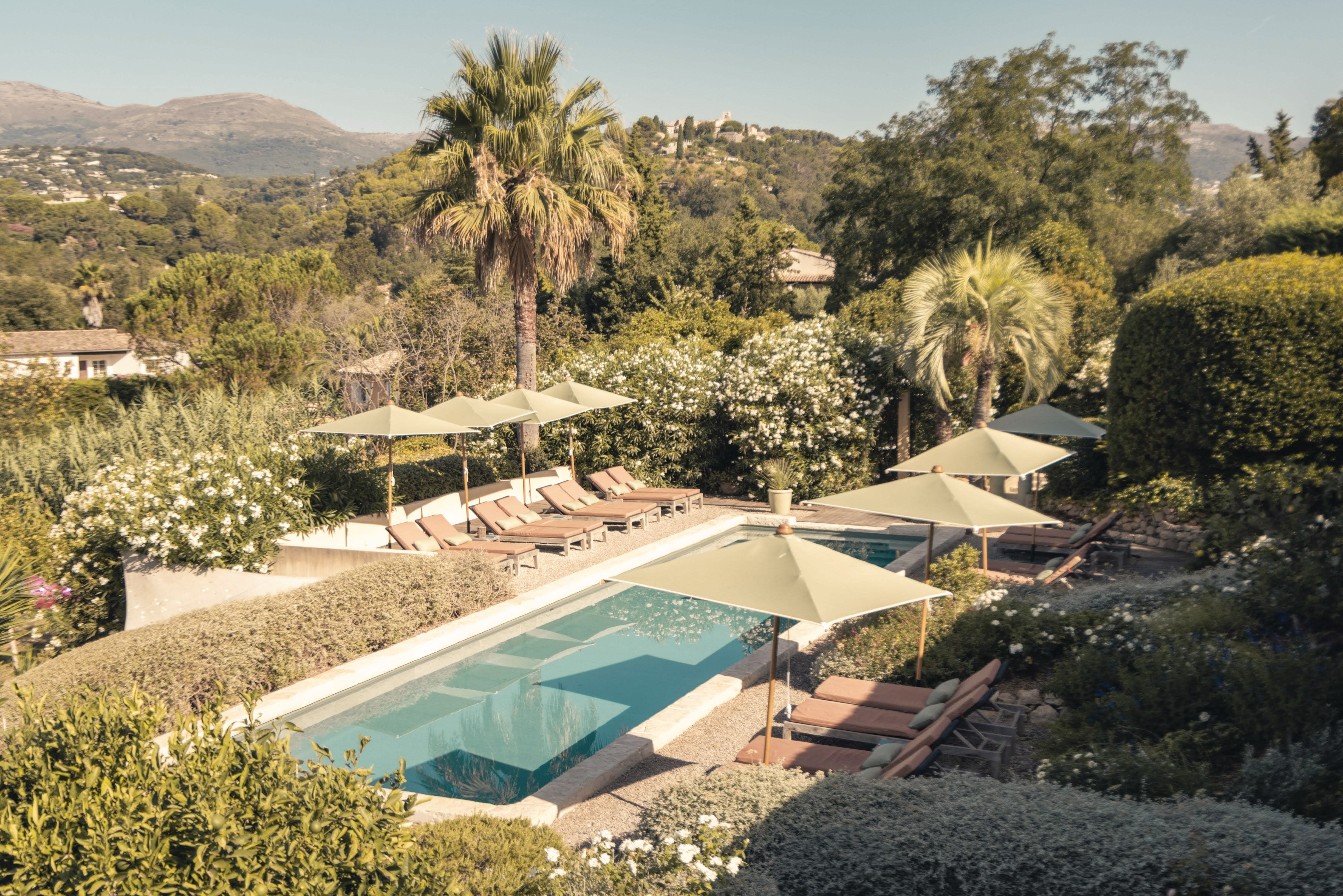 Outdoor pool surrounded by sun loungers with umbrellas, lush greenery, palm trees, and hills in the background