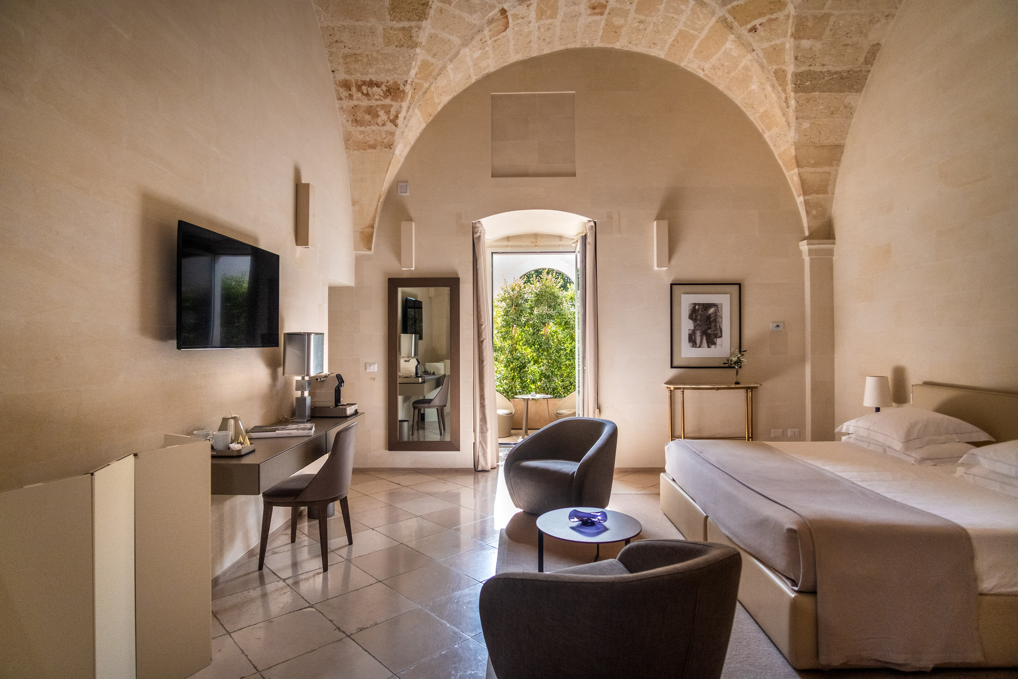 Bright hotel room with arched stone ceiling, king bed, two armchairs with a small table, a desk with chair, and an open door leading to a leafy outdoor space.