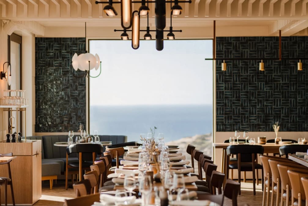 Elegant restaurant interior with long wooden tables set for dining and a large window offering a view of the sea.