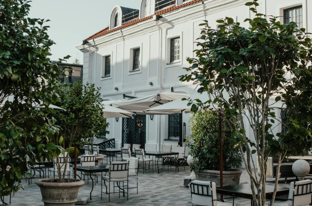 Outdoor courtyard with black tables and white cushioned chairs, large potted plants, umbrellas, and a white building in the background.