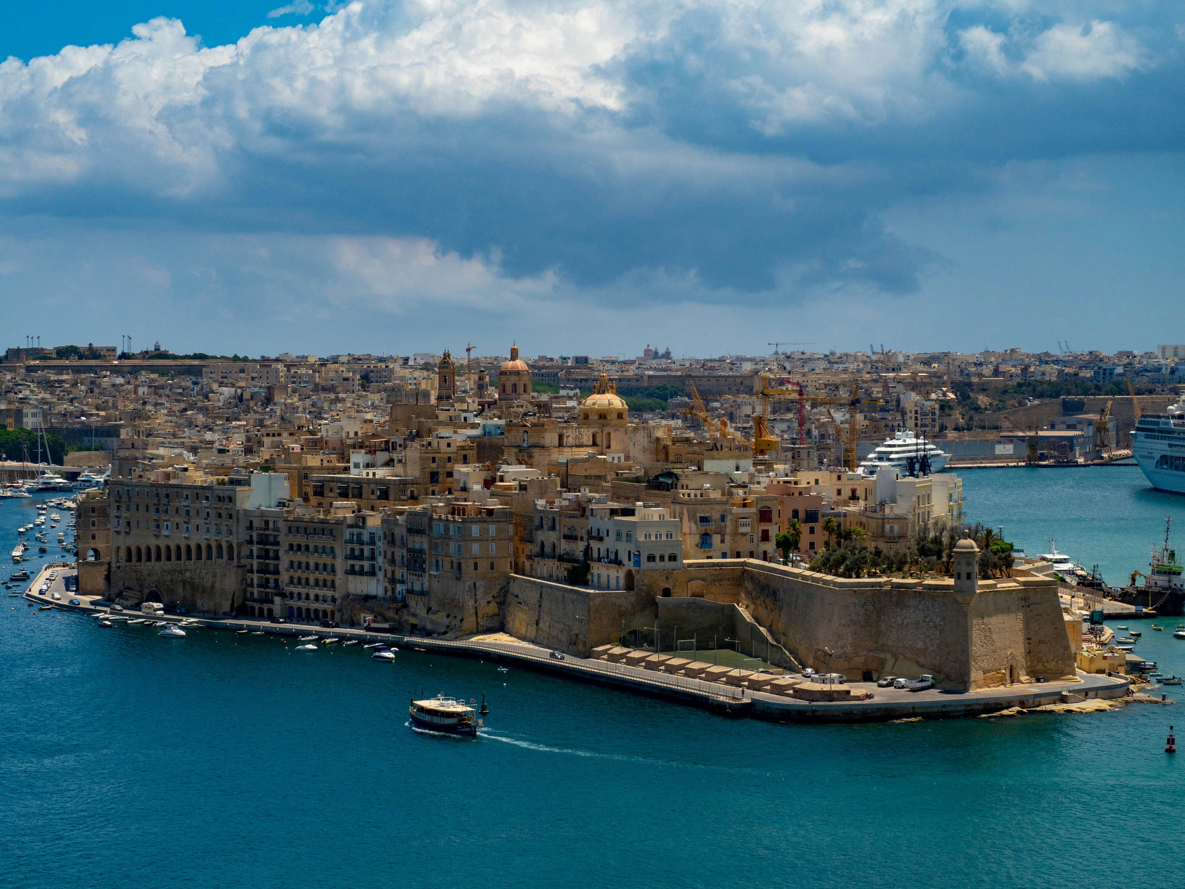 Aerial view of Valletta, Malta, showing historic stone buildings, fortifications, and boats on clear blue harbor waters under a partly cloudy sky