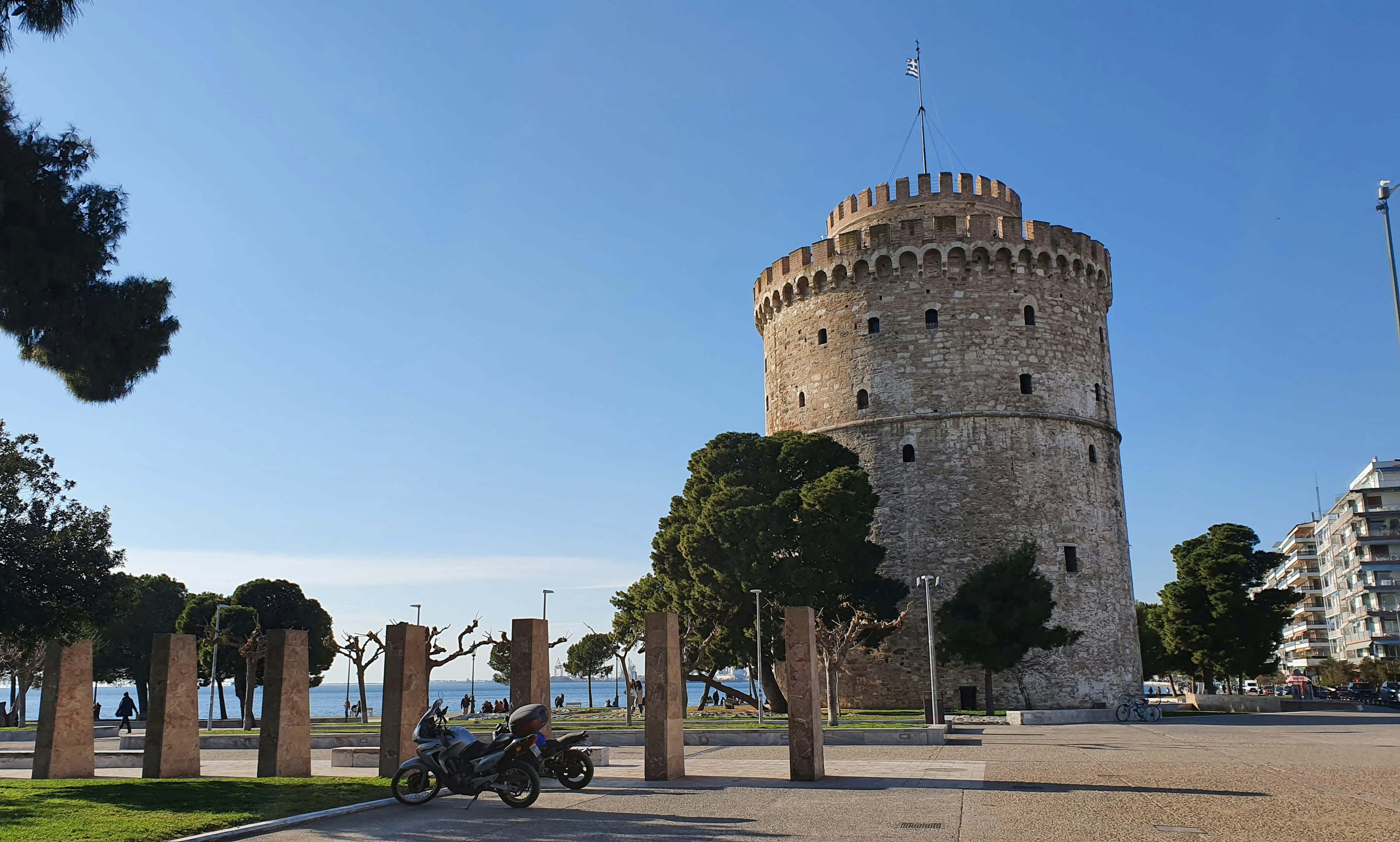 Stone tower with battlements next to a park with trees and stone pillars, motorcycles parked on a paved area, and the sea in the background under a clear sky.