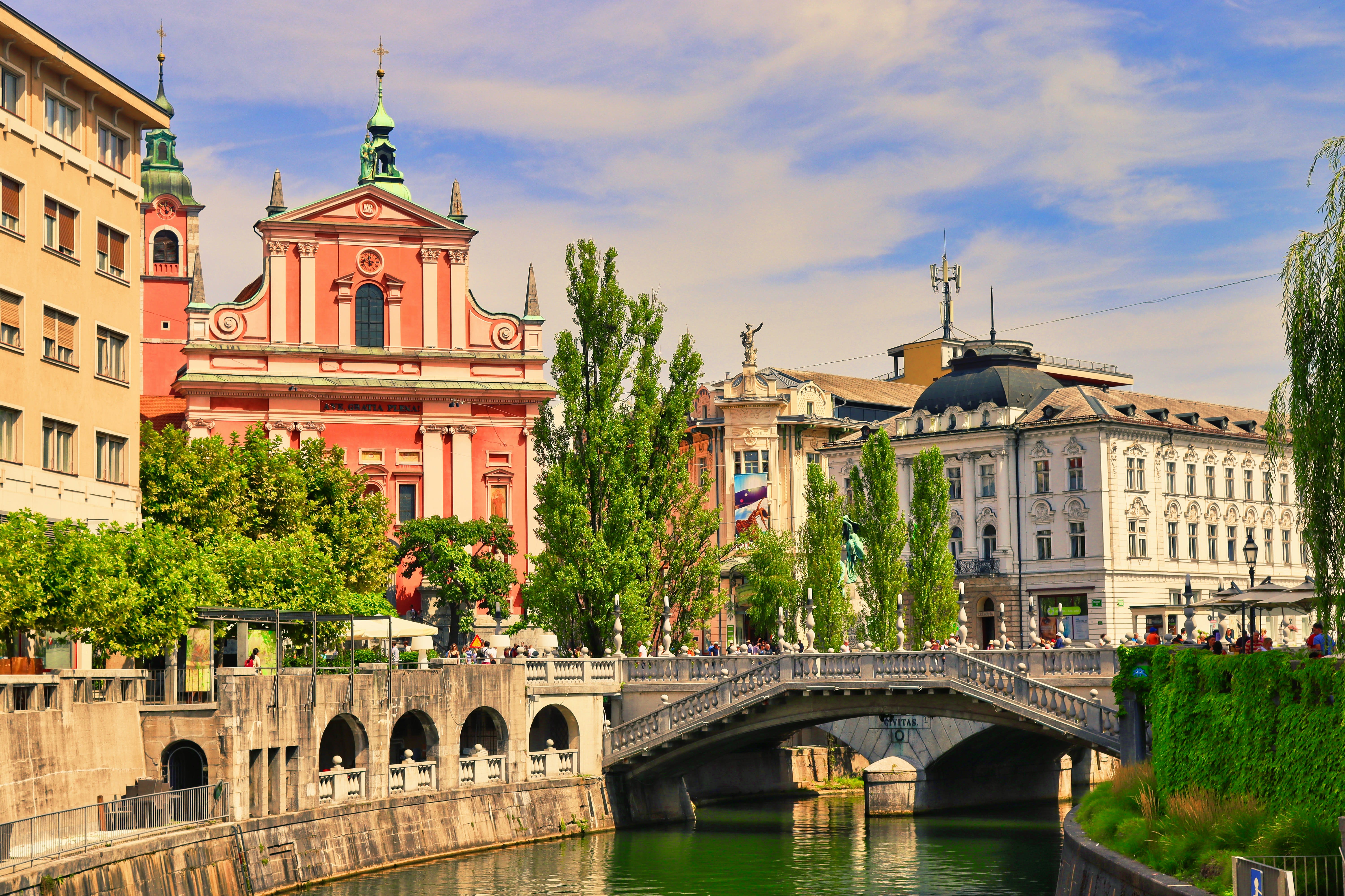 River with a stone bridge, lined by historic buildings and leafy trees, with a vibrant pink church facade in the background.
