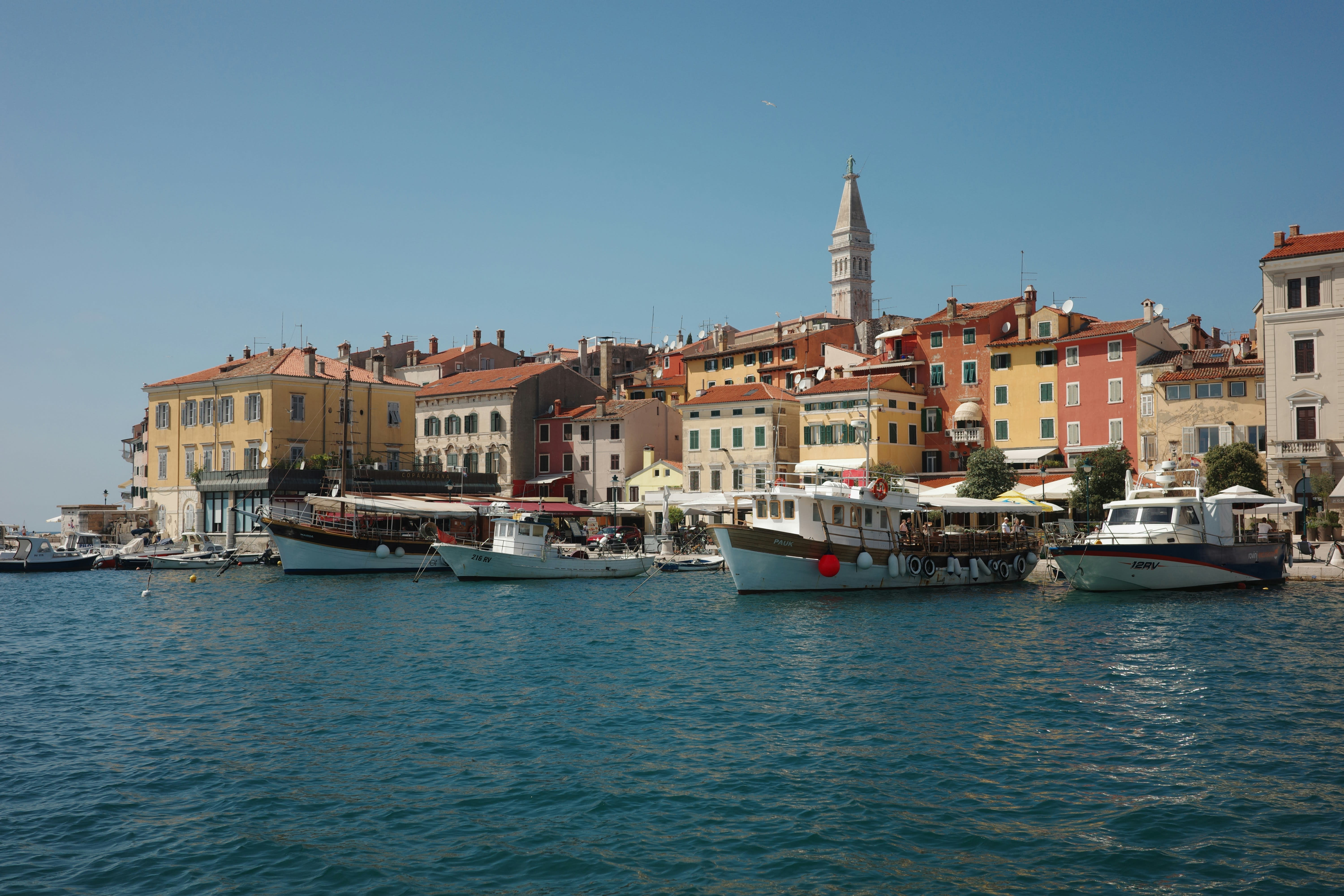Colorful coastal town with boats docked in a harbor and a tall church tower rising above terracotta-roofed buildings.