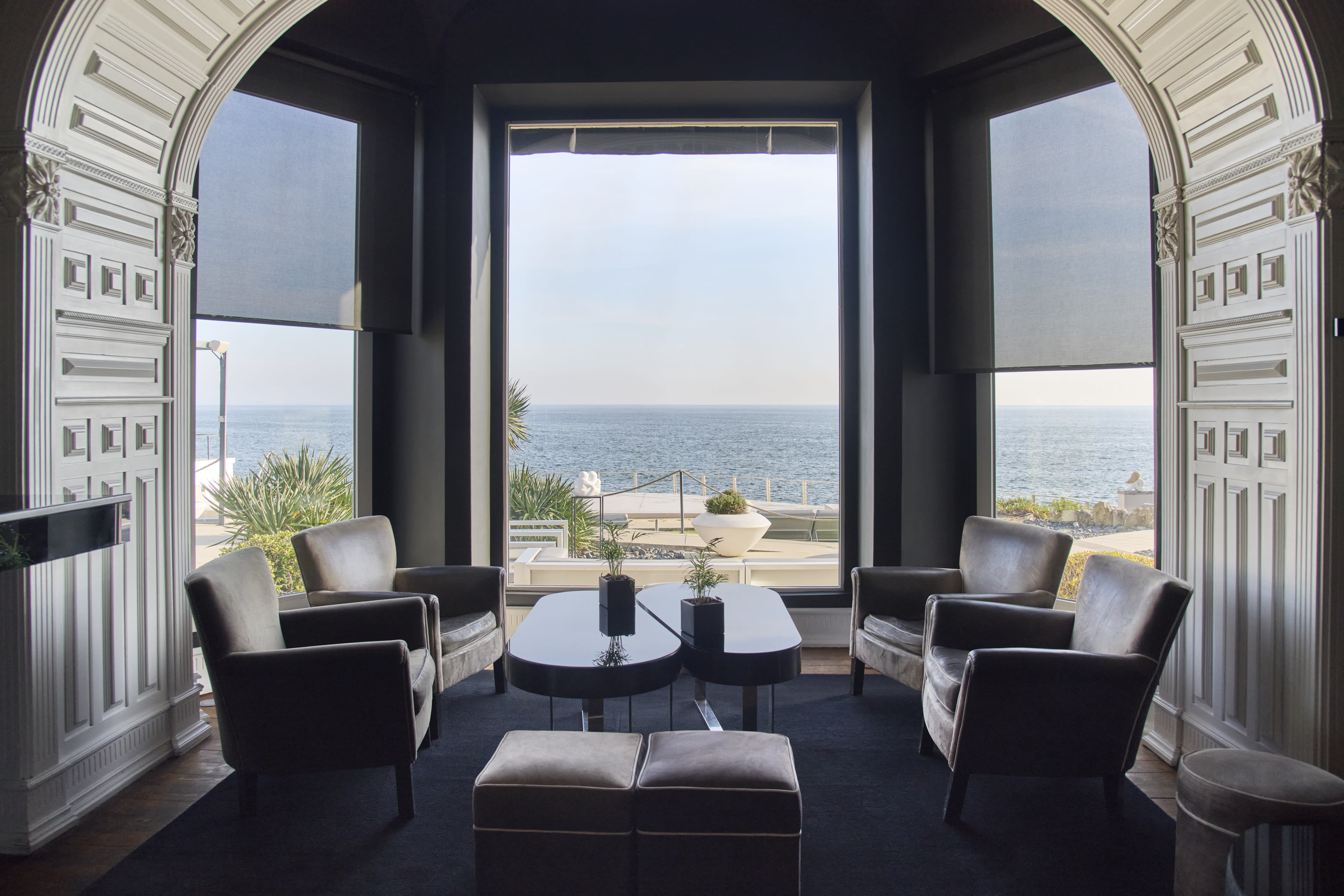Cozy lounge with leather armchairs arranged around two dark tables, large windows framing a view of the sea and coastal plants.