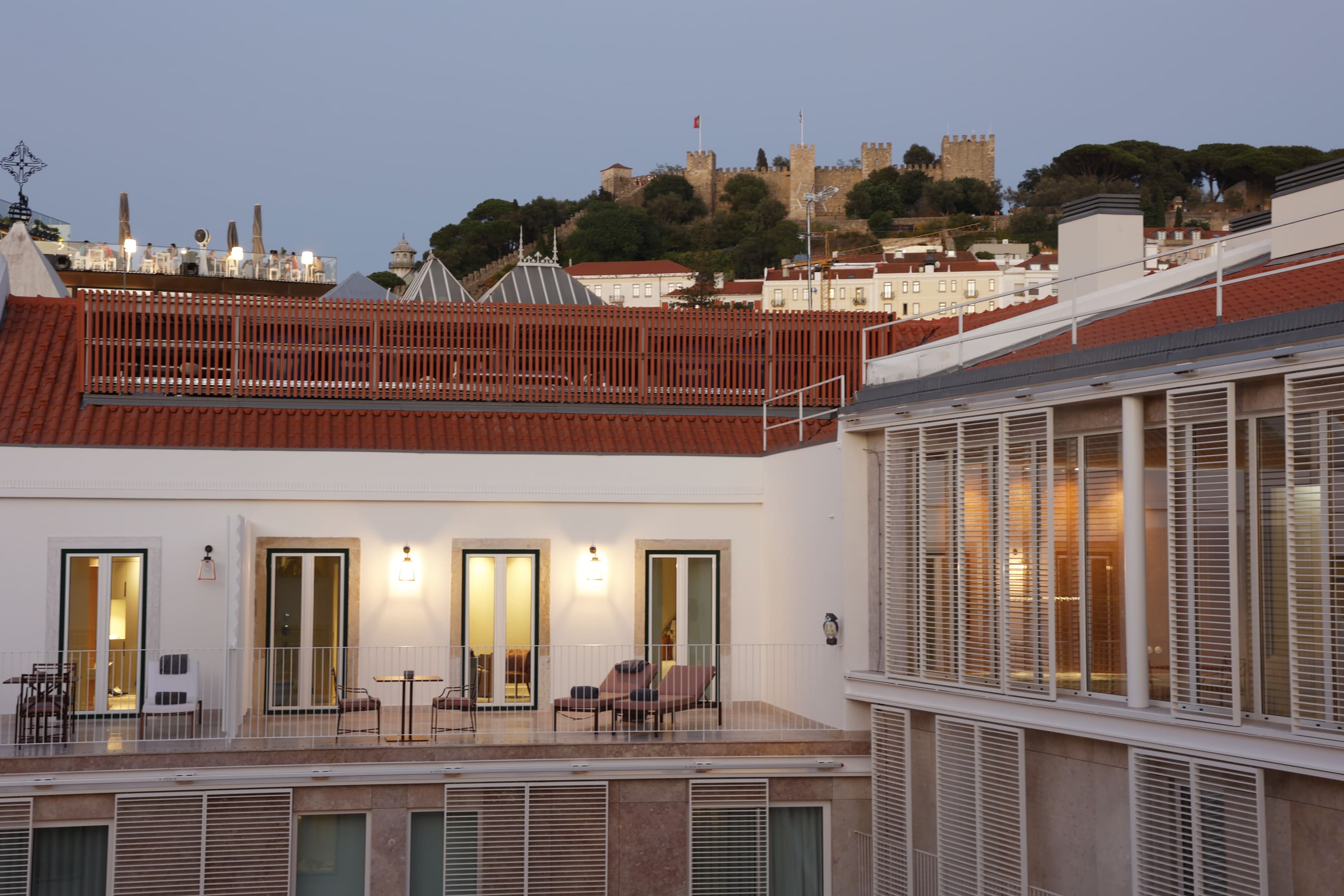 Hotel terrace with chairs, tables, and loungers lit by warm lights, with a castle on a hill visible in the background at dusk.