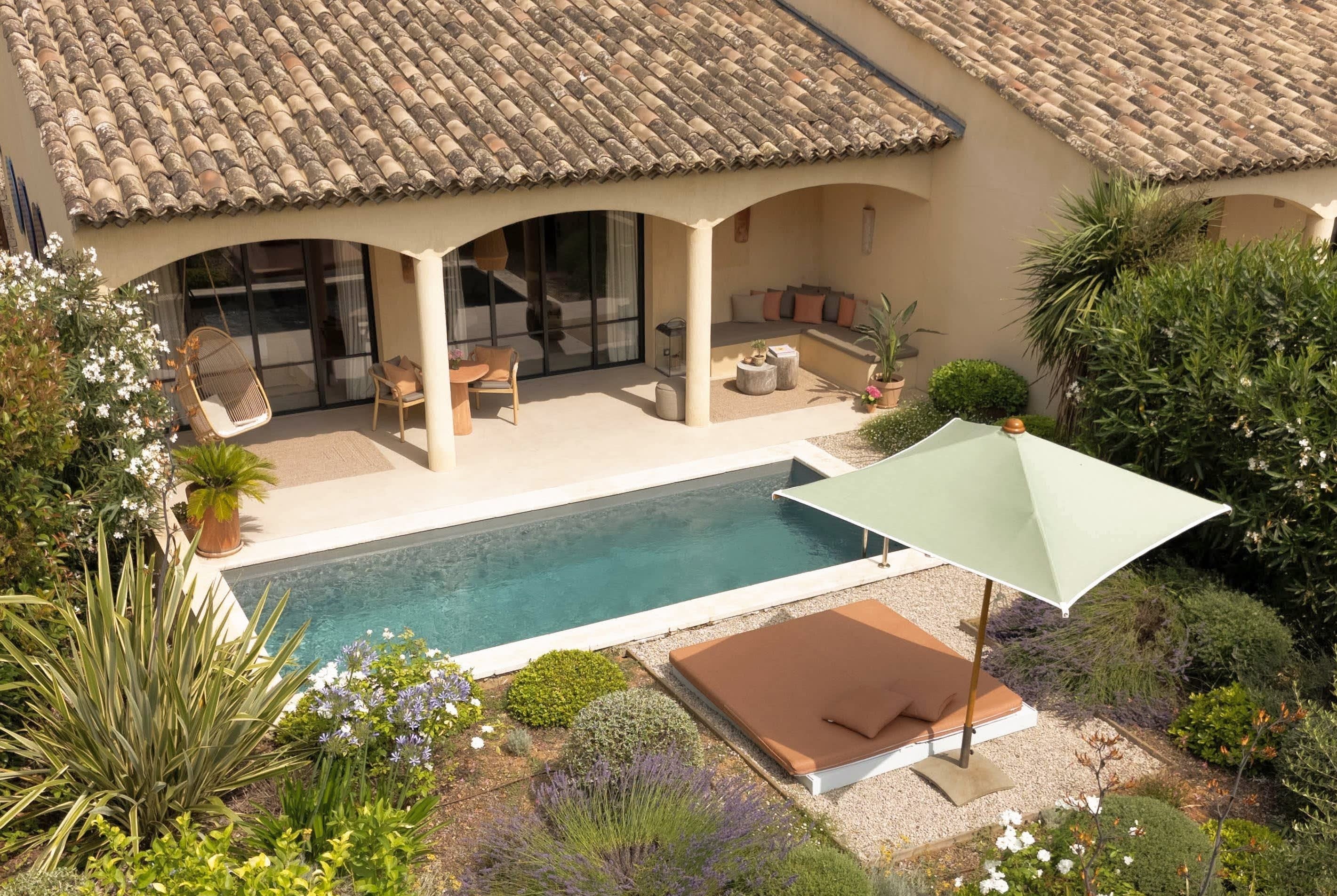 Garden with flowering shrubs and plants surrounding a rectangular pool, adjacent to a covered patio with seating and a large sunbed with a green umbrella.