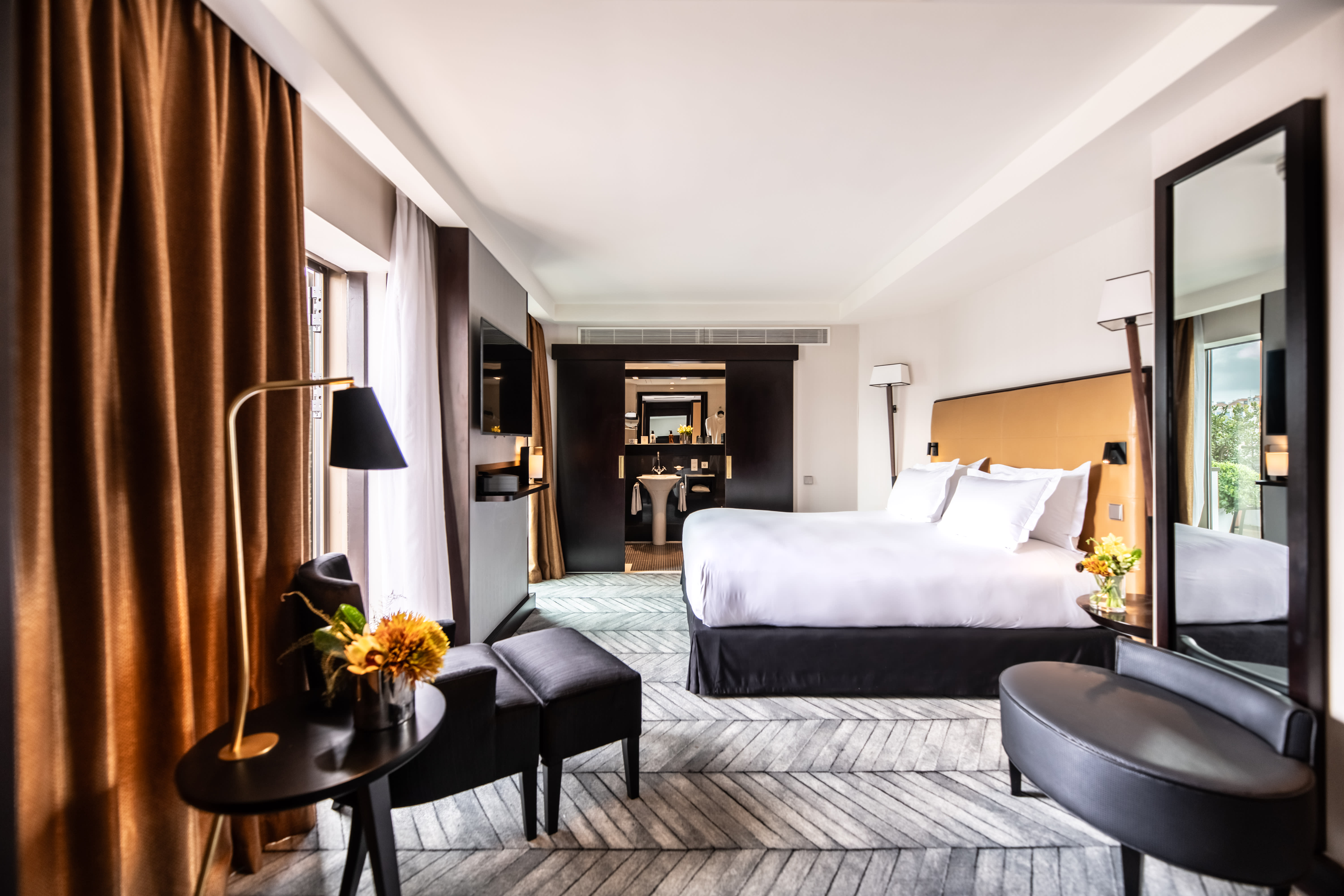 A modern hotel bedroom with a large bed, black and gold accents, and a seating area by a window with brown curtains.