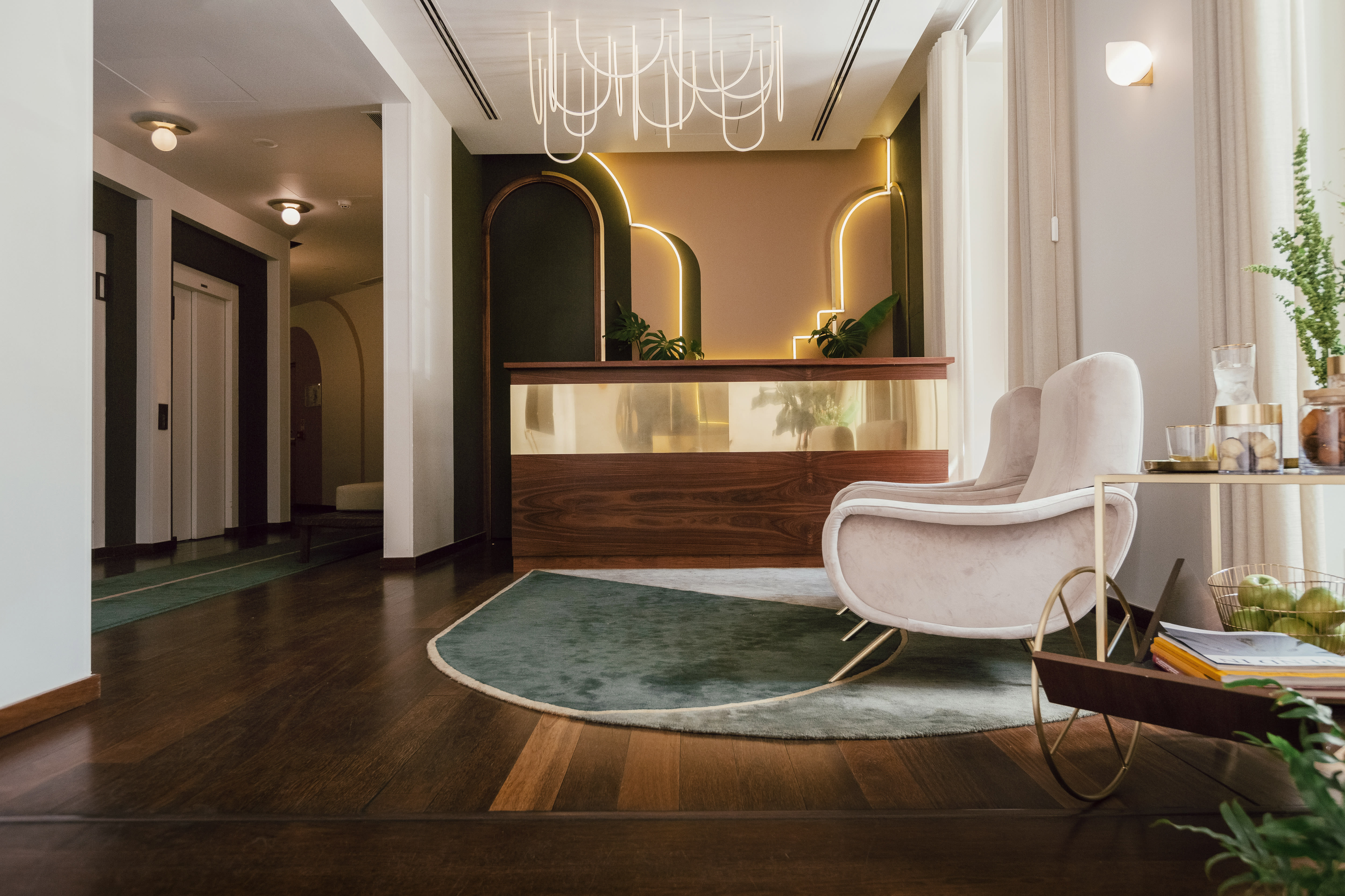 A stylish hotel lobby with a wooden reception desk, curved soft pink chairs, green rug, and elegant wall lighting.