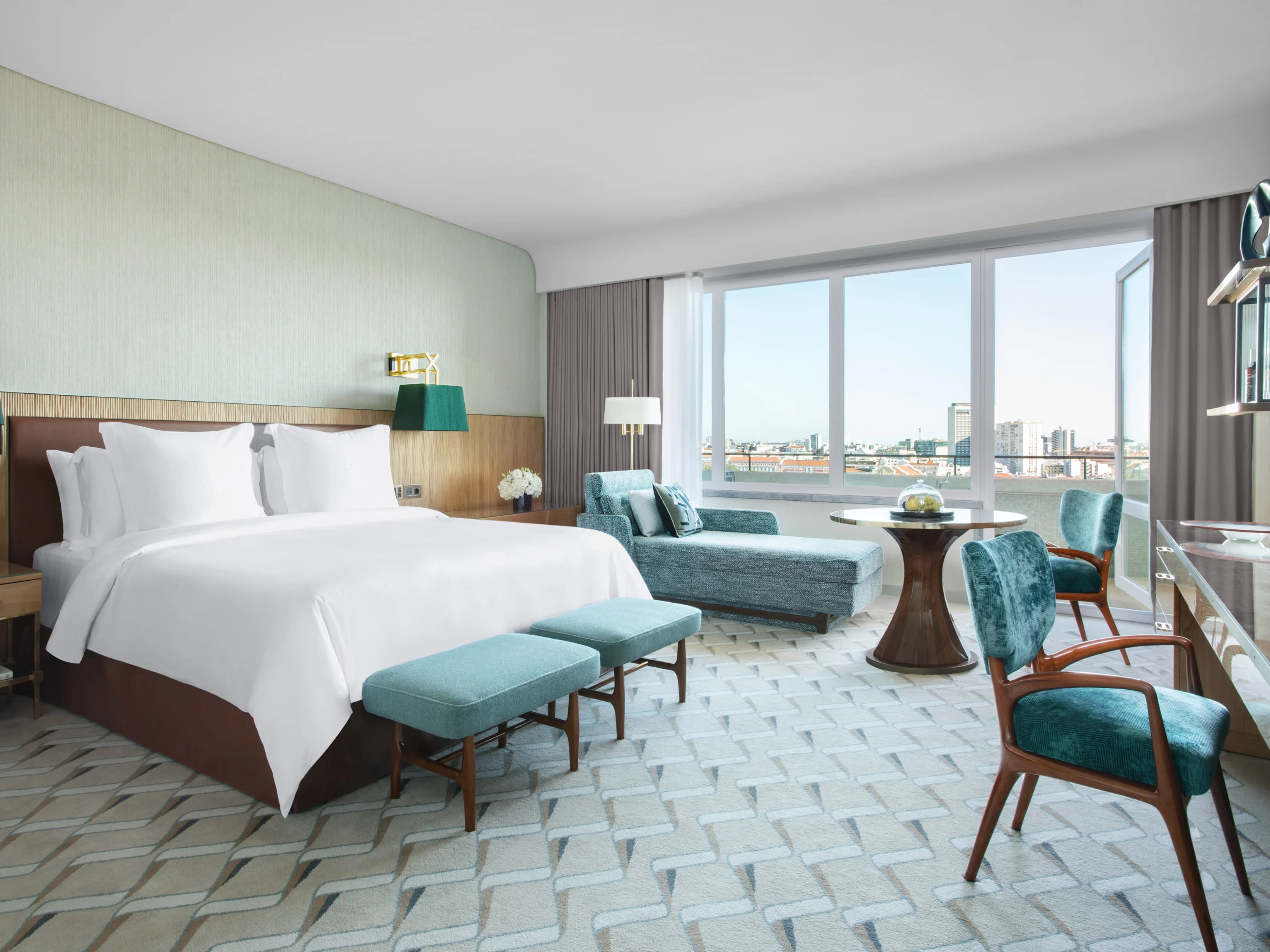 A hotel room with light grey carpet, white linens, and greenish teal accents like chairs, lamps and chaise lounge