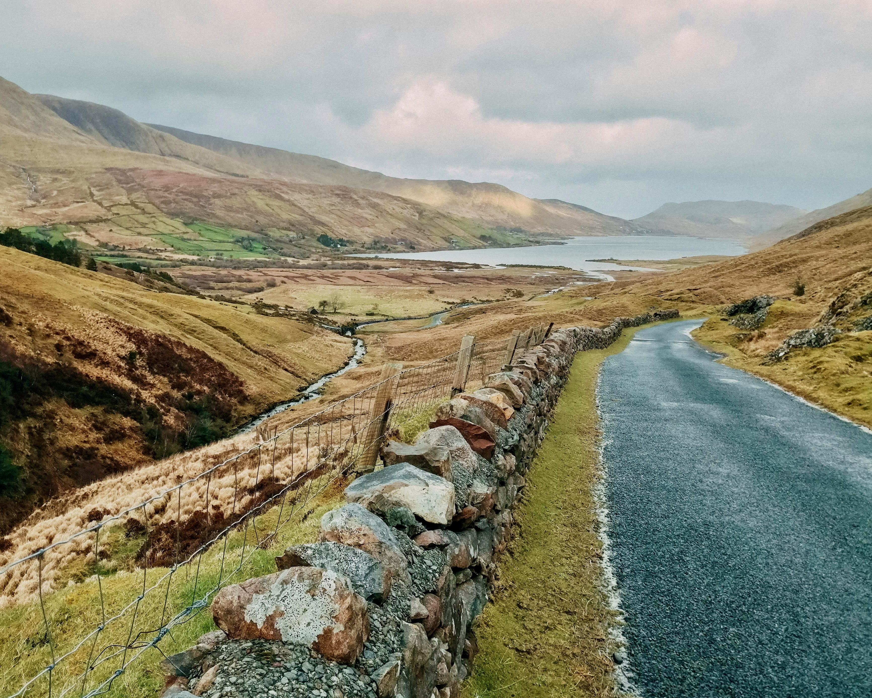 Narrow road with stone wall in mountainous terrain and lake in the distance under cloudy daytime skies