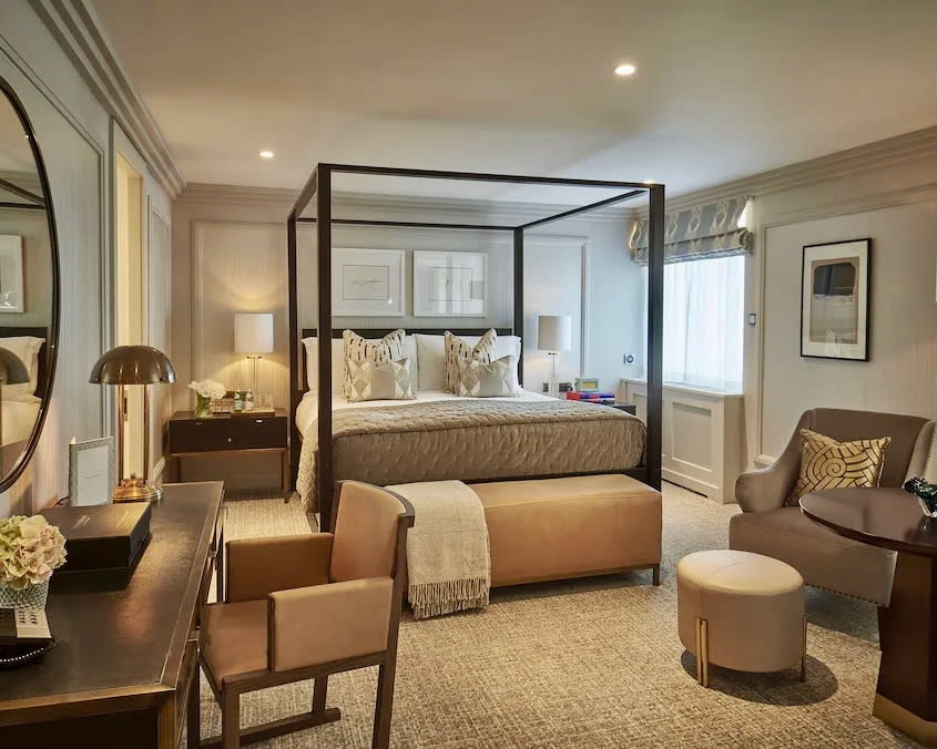 earth toned hotel room with black four poster bed, tan bench and desk chair, and beige sitting chair and bedding