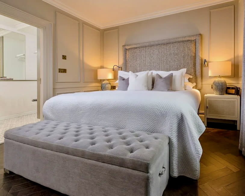 white walled hotel room with beige headboard and bench, white linens, and wood floors