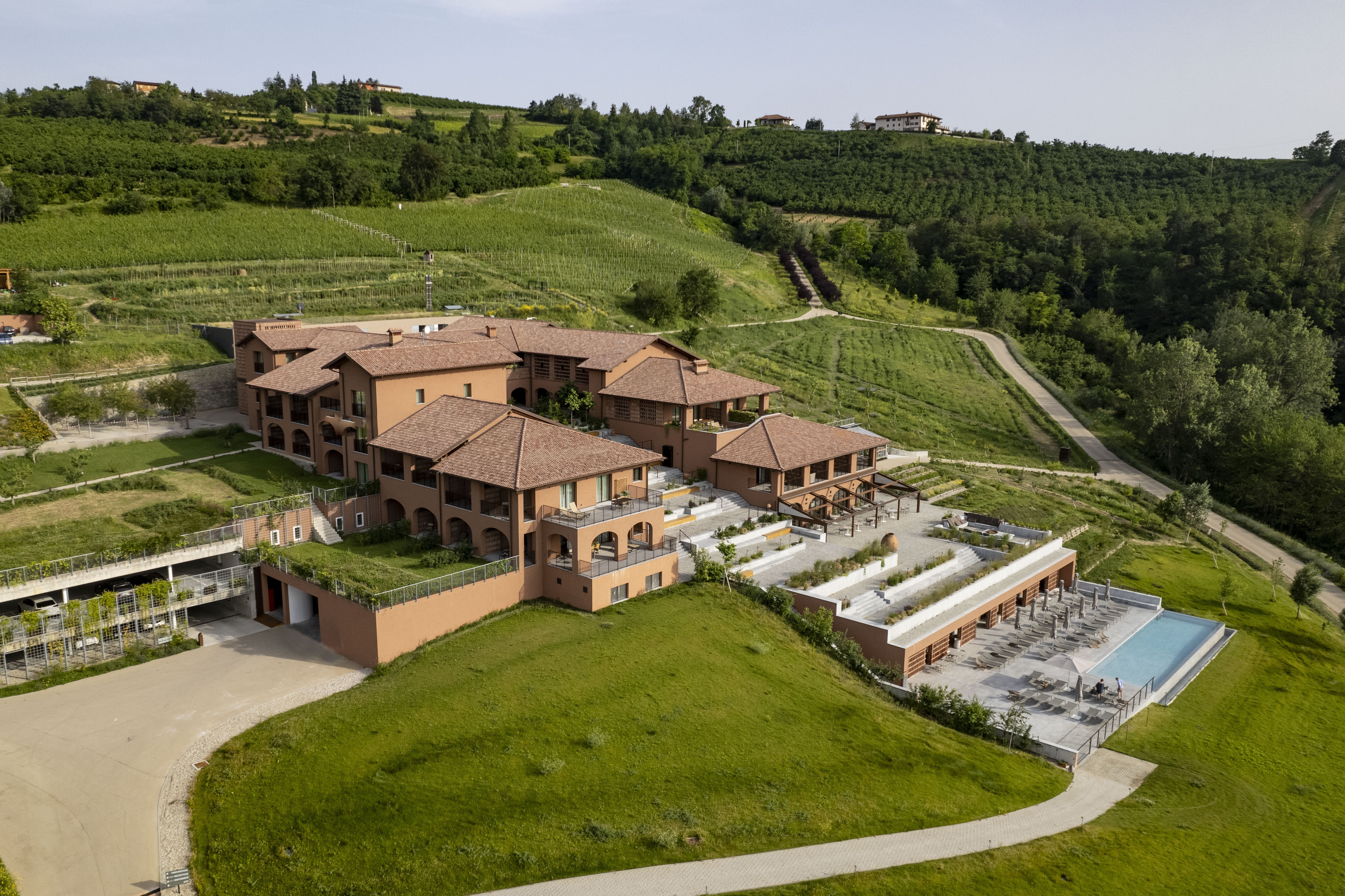 An aerial view of a large terracotta-roofed hotel surroudned by green rolling hills with terraces, a pool, and vineyards.