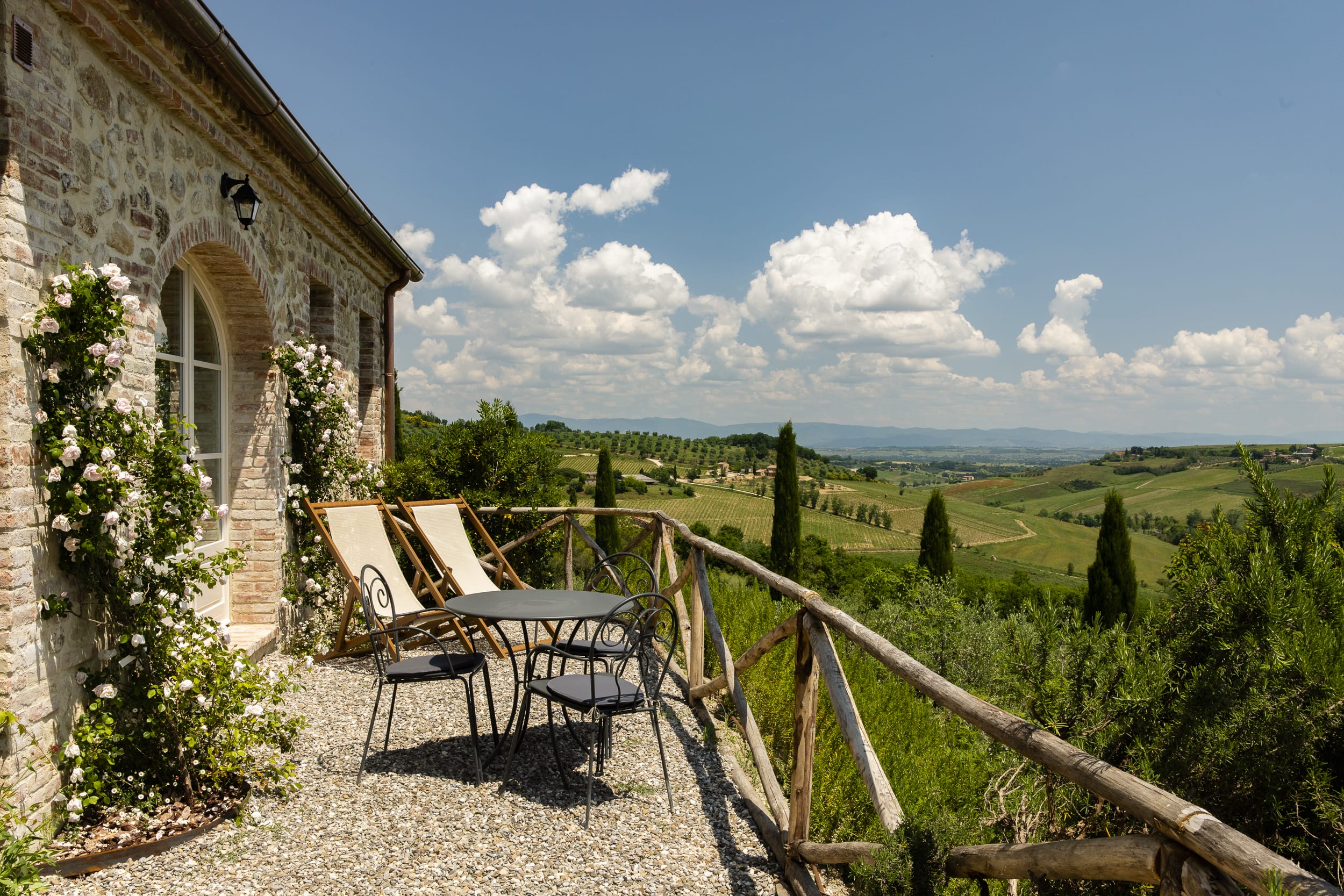 Stone villa terrace with table and chairs overlooking rolling green Tuscan hills under a partly cloudy blue sky.