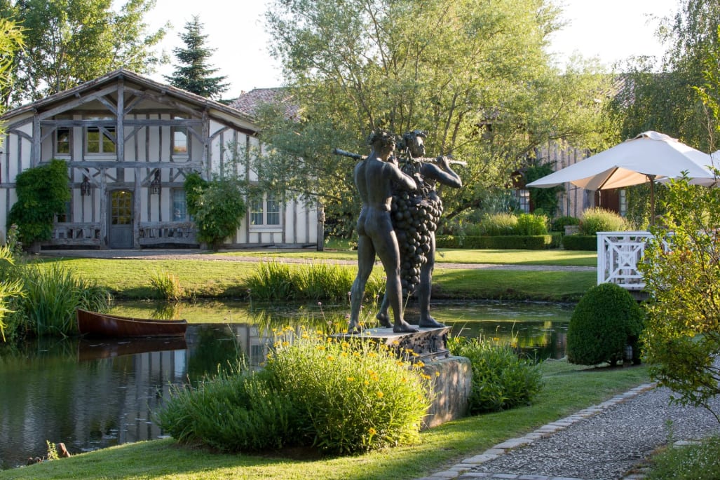 Bronze statue of two men playing flutes surrounded by lush gardens and a pond with a wooden boat, with a rustic half-timbered building and white umbrella in the background