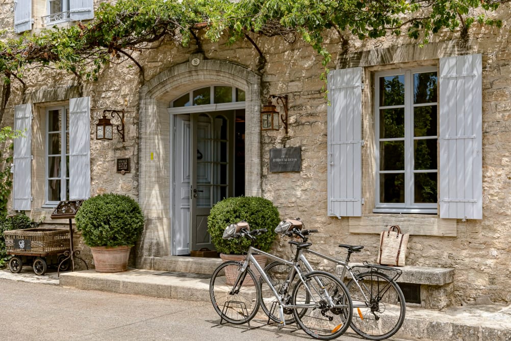 Stone facade of a charming hotel entrance with light gray shutters, potted plants, and two bicycles parked outside under a vine-covered trellis.