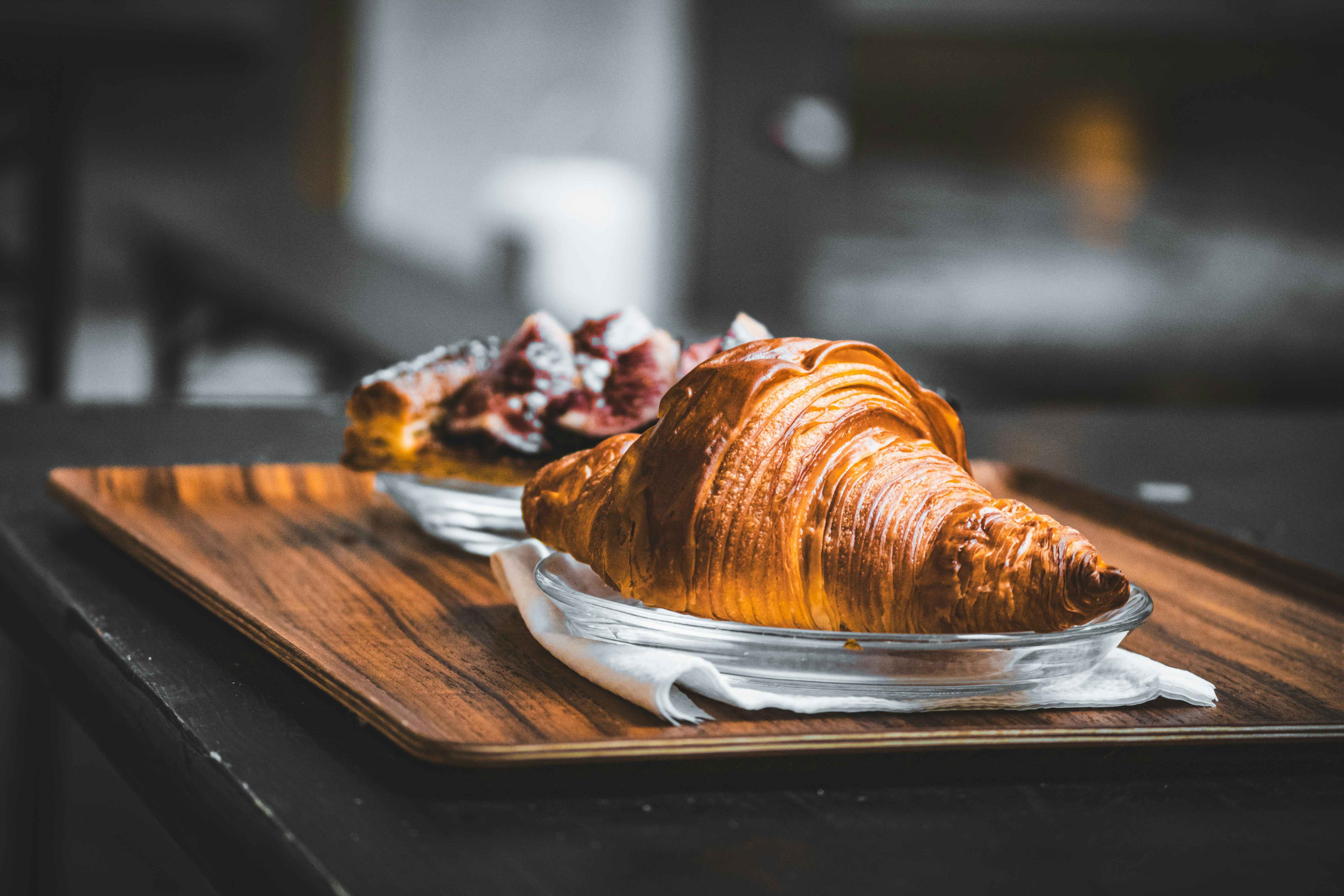 Buttery, flaky croissant served on a clear glass plate with a blurred background of other pastries on a wooden tray.