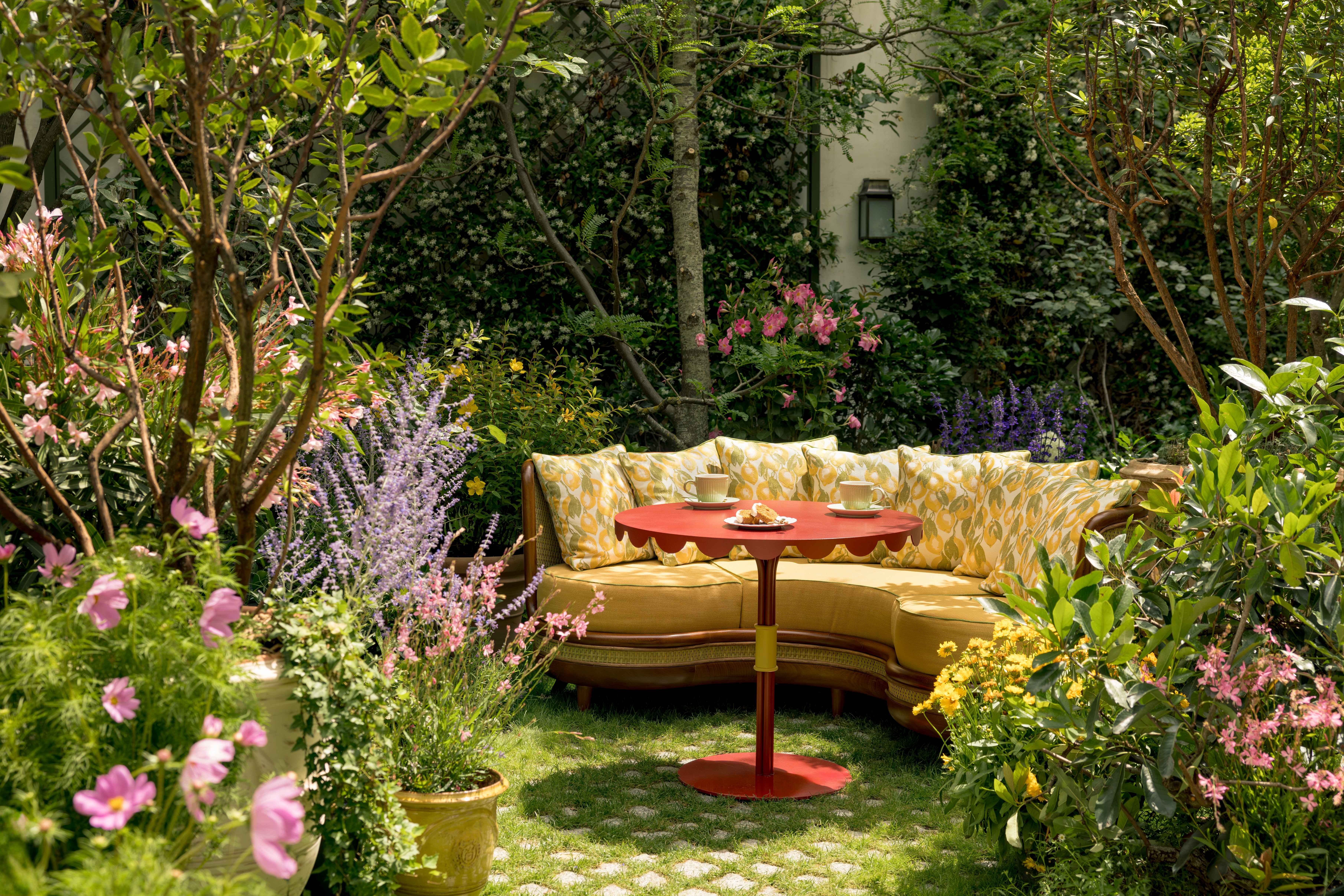 Outdoor garden seating with a curved yellow sofa and a red scalloped-edge table surrounded by lush greenery and colorful flowers.