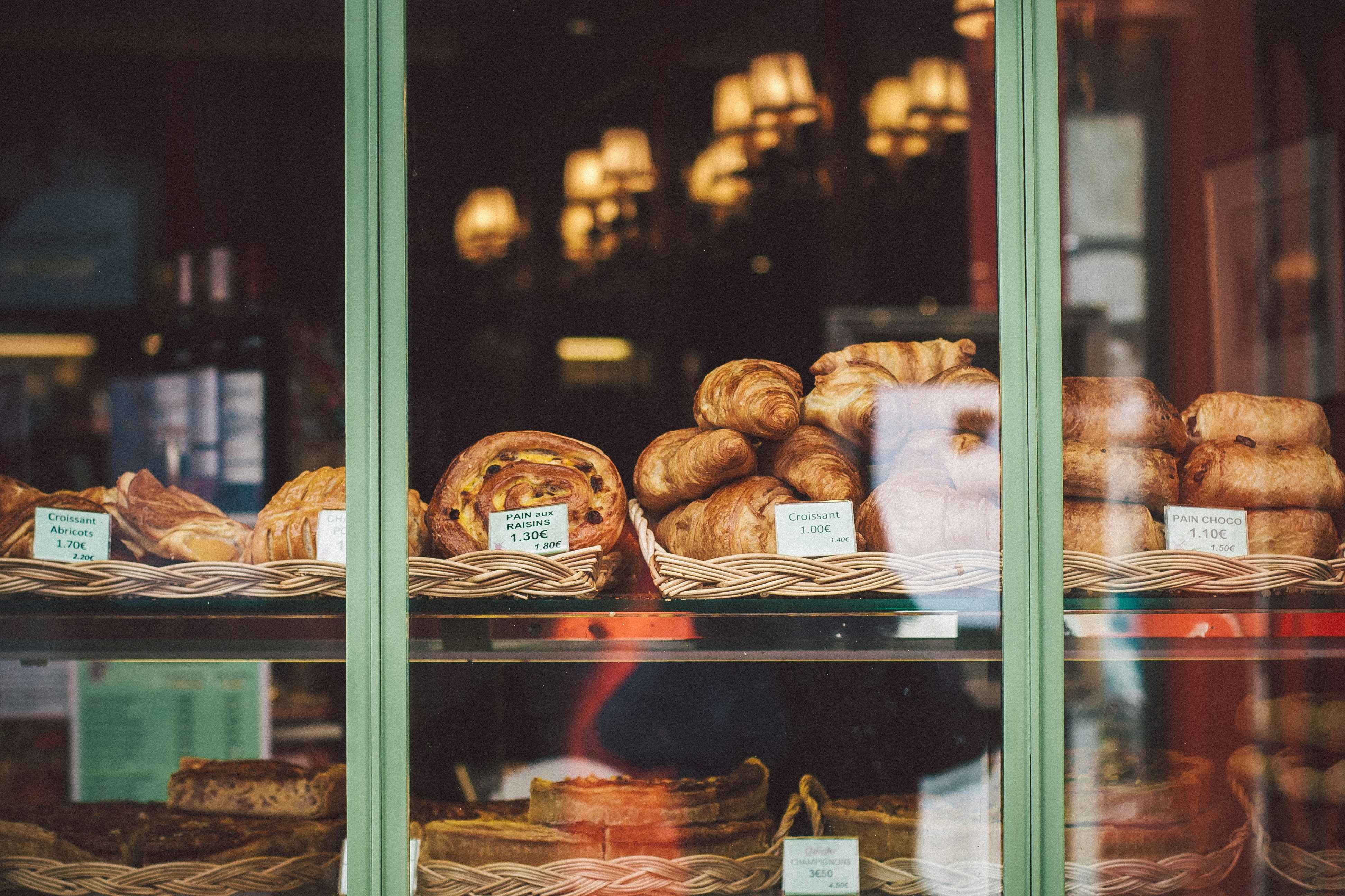 Pastries including croissants and pain au chocolat displayed in baskets behind a green-framed glass window at a bakery.