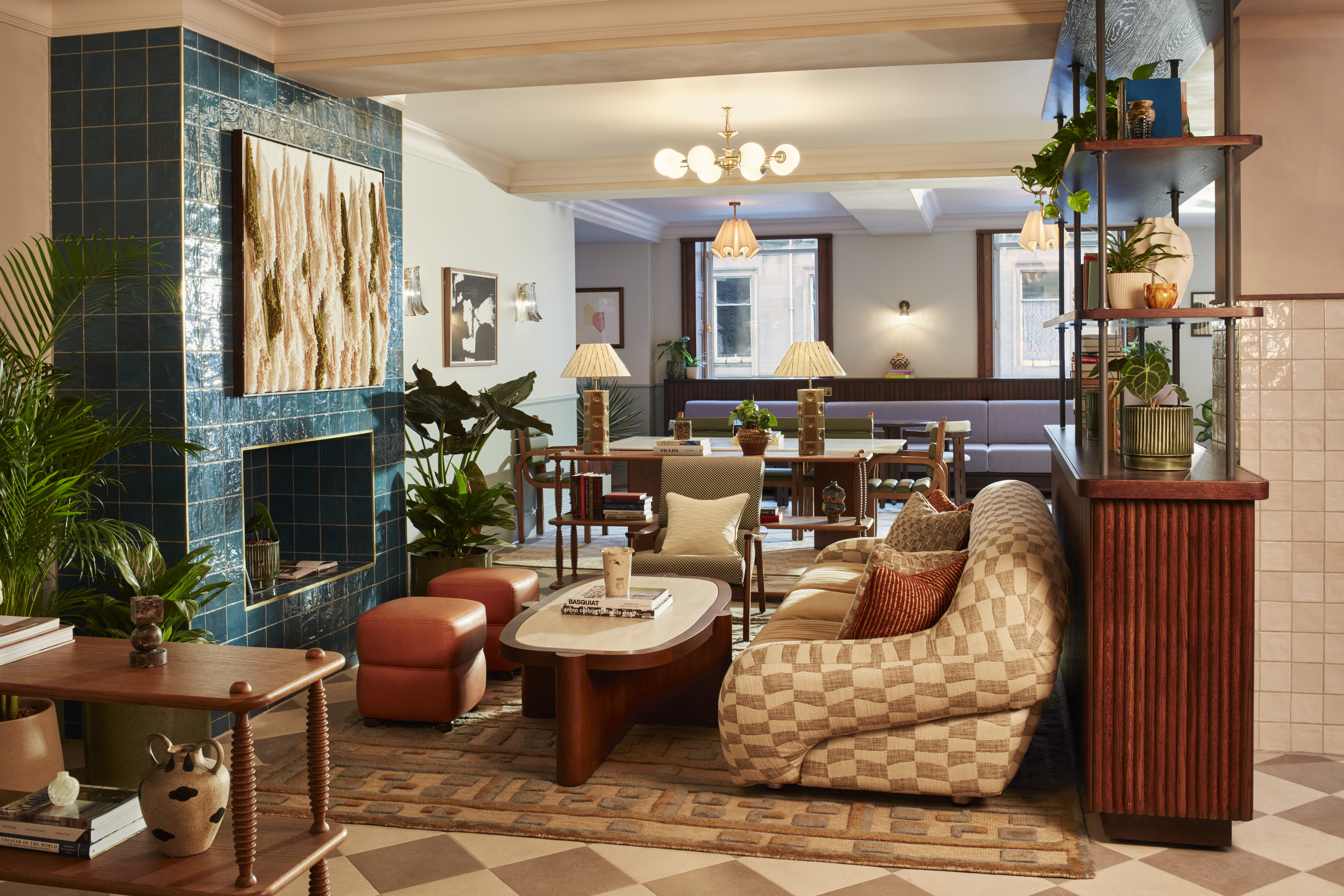 Cozy hotel lobby featuring a checkered sofa, teal tiled fireplace, vintage wooden furniture, and lush plants, creating a warm and inviting atmosphere.